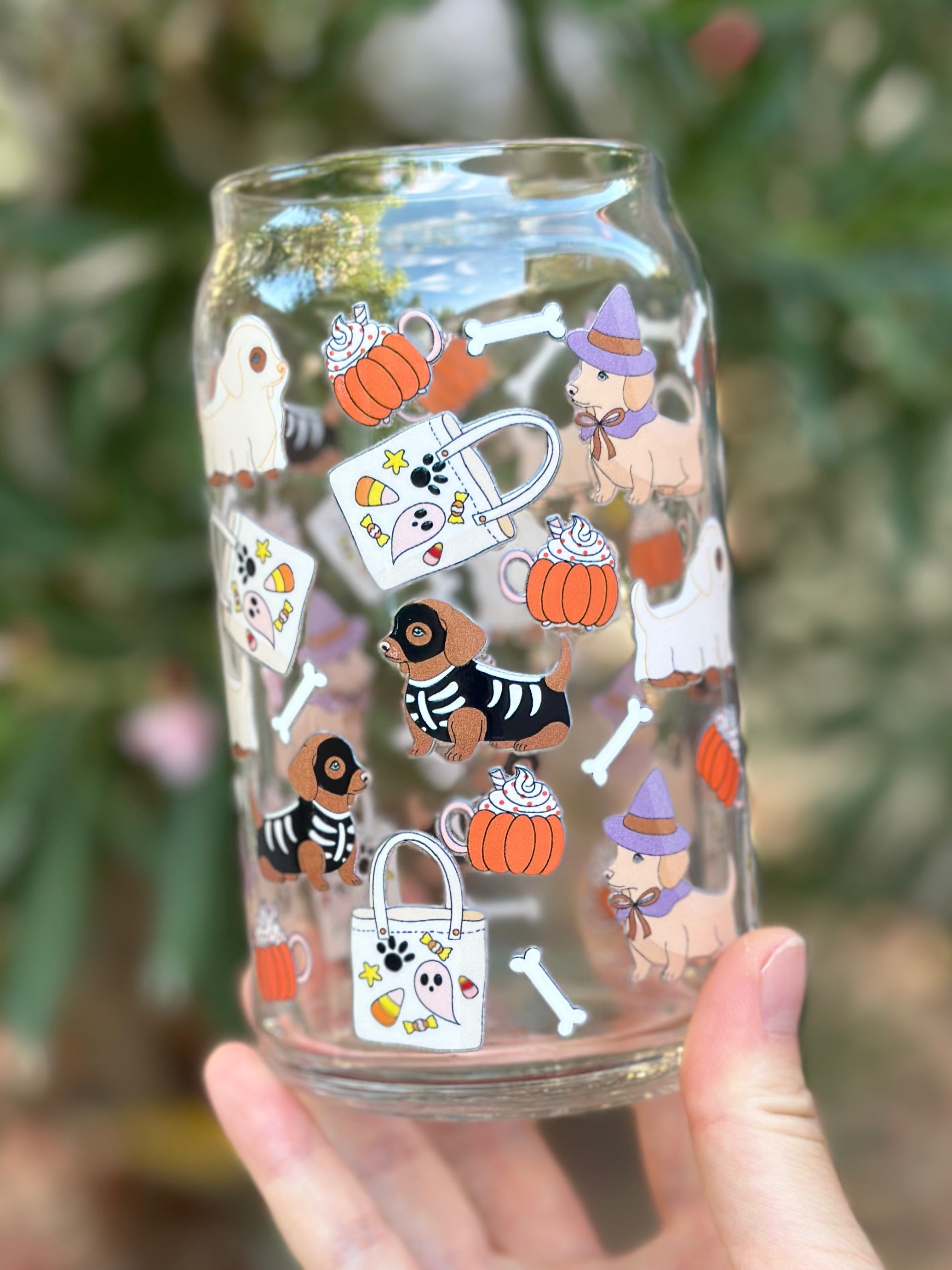 Clear glass can cup decorated with dachshunds in Halloween costumes, including a witch hat, skeleton outfit, and ghost costume, surrounded by pumpkins, candy, bones, and tote bags.