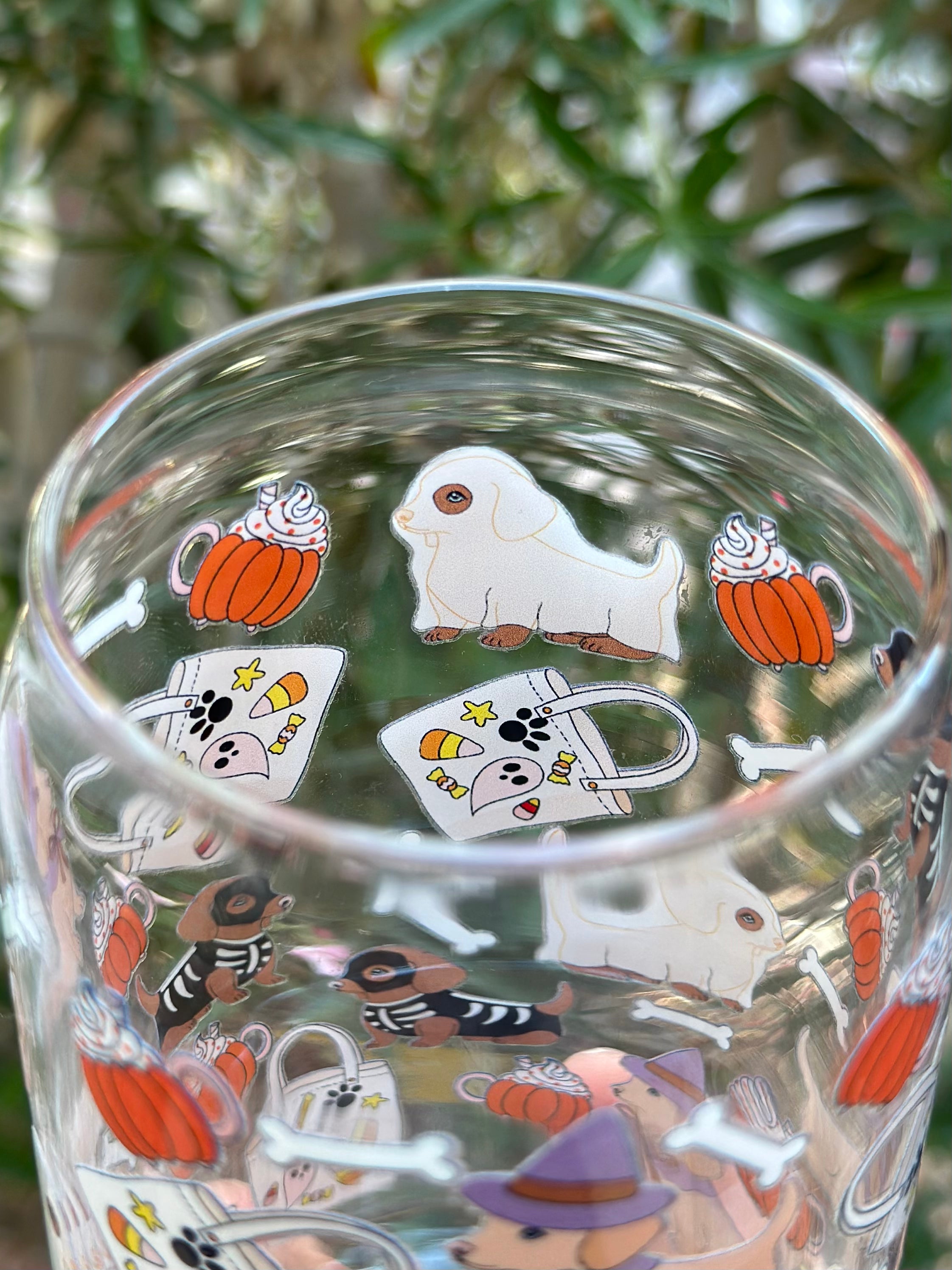 Inside view of glass can cup showing a dachshund in a ghost costume alongside pumpkins, candy mugs, tote bags, and bones.