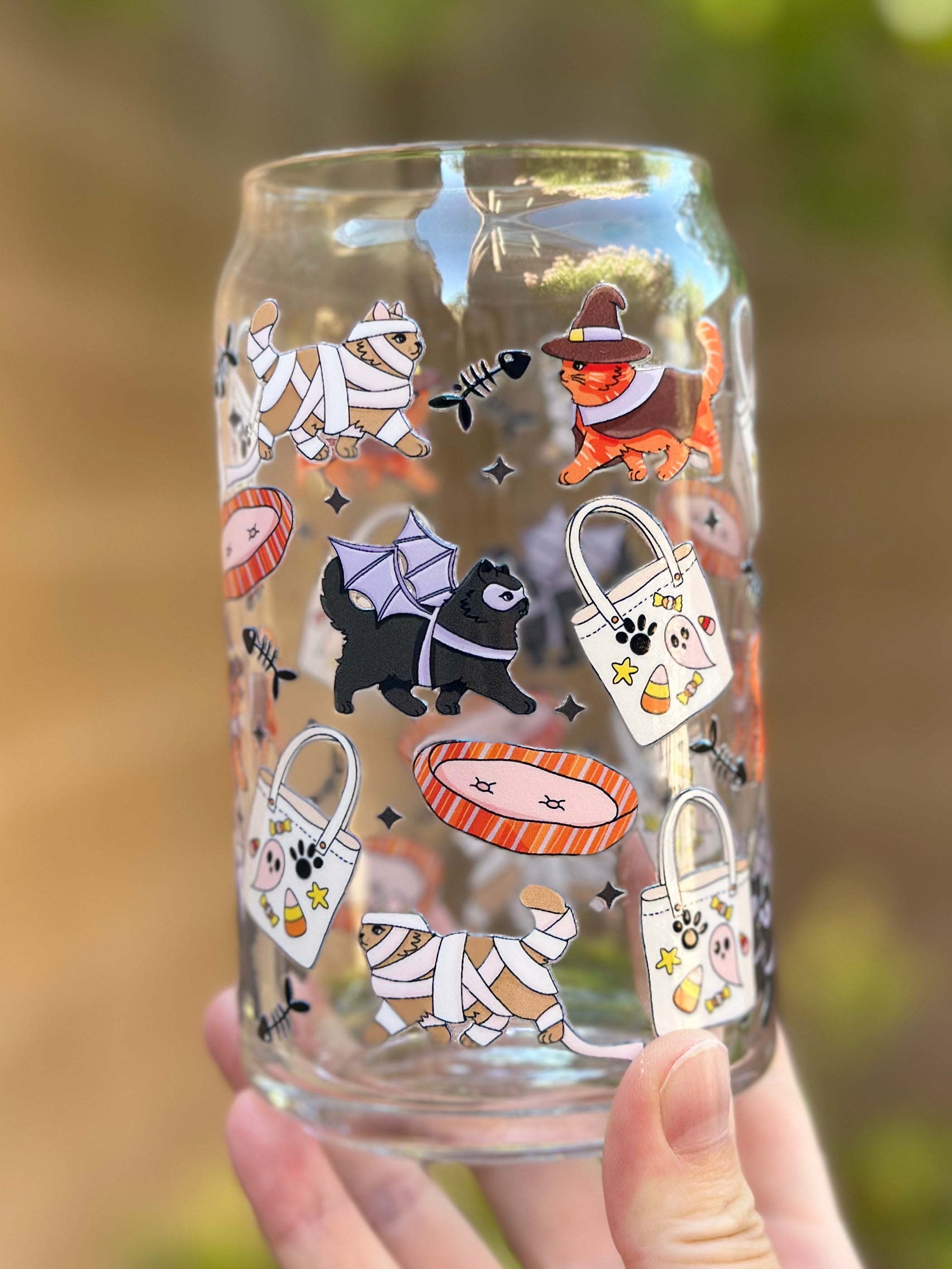 Clear glass can cup decorated with cute Halloween cats and dogs in costumes, including a witch cat, mummy cats, and a bat-wing dog, surrounded by candy bags, fish bones, and pet beds.