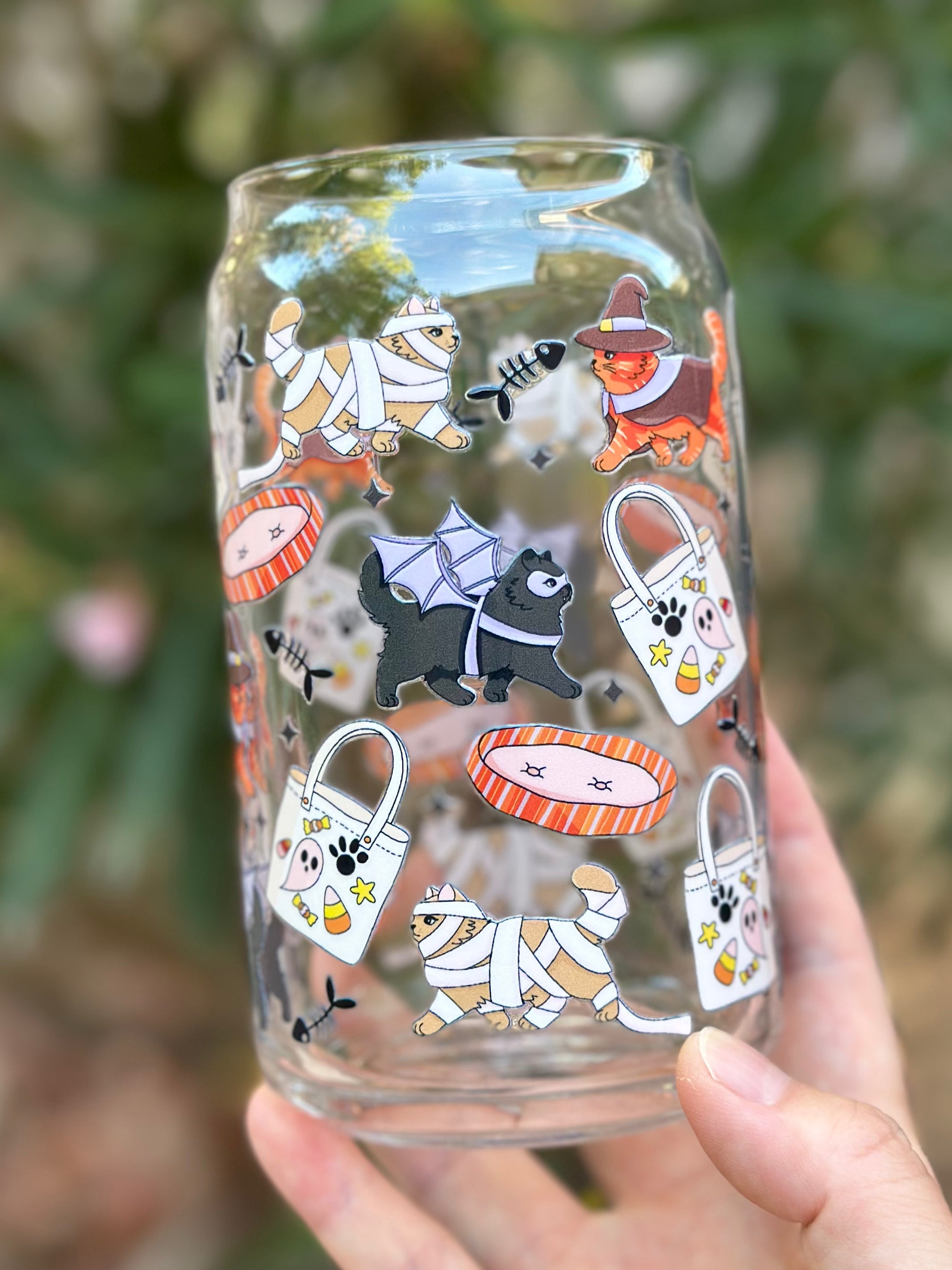 Clear glass can cup decorated with cute Halloween cats and dogs in costumes, including a witch cat, mummy cats, and a bat-wing dog, surrounded by candy bags, fish bones, and pet beds.