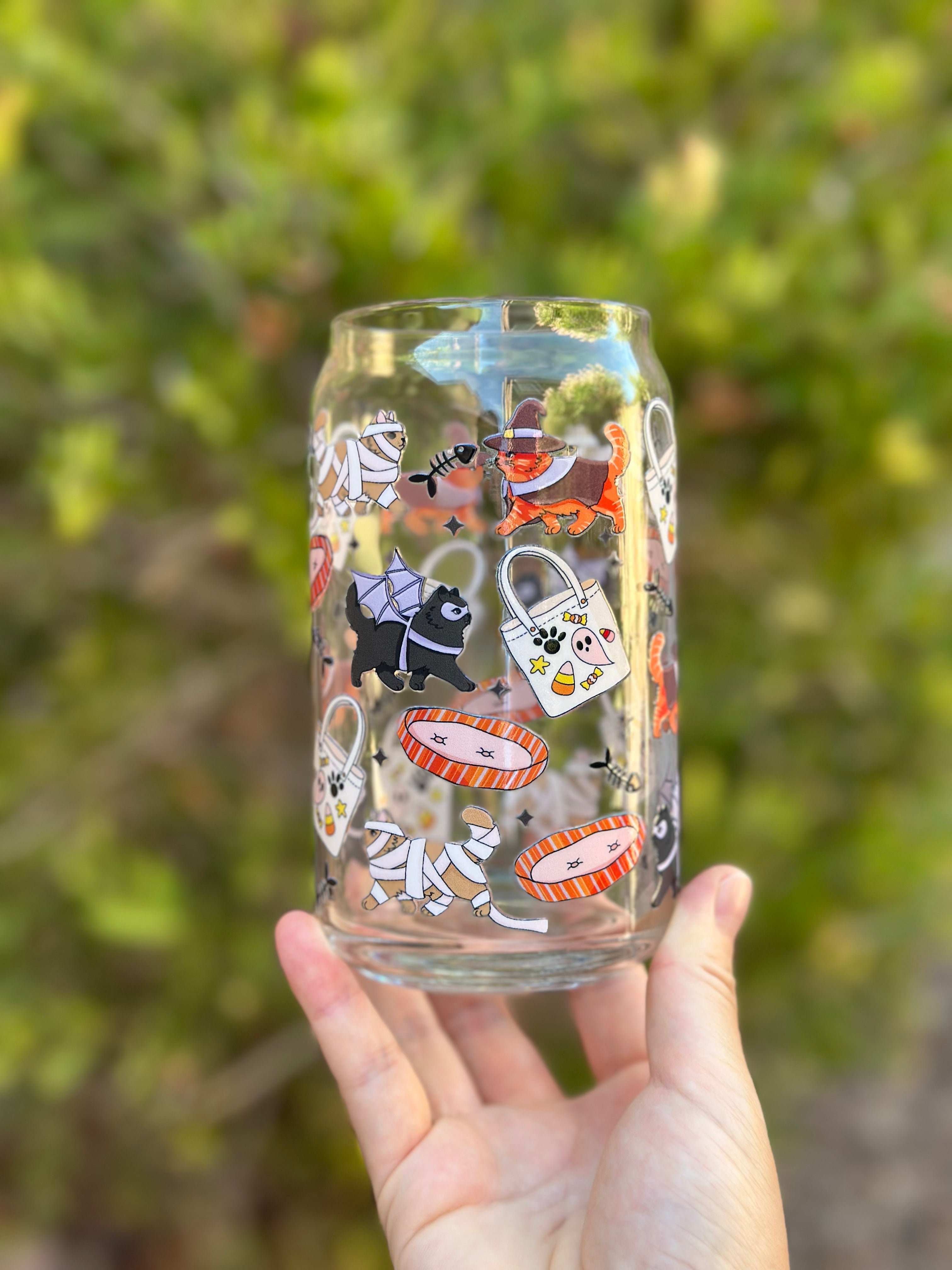 Clear glass can cup decorated with cute Halloween cats and dogs in costumes, including a witch cat, mummy cats, and a bat-wing dog, surrounded by candy bags, fish bones, and pet beds.