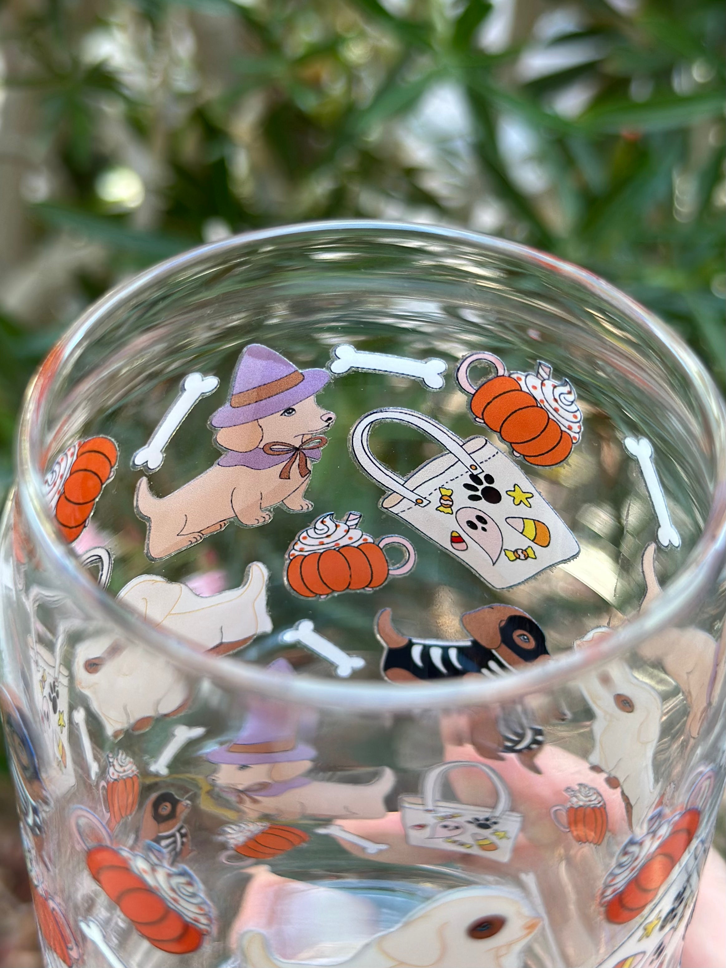 Close-up of dachshund in a purple witch hat with surrounding pumpkins, candy mugs, tote bags, and bones on a clear glass can cup.