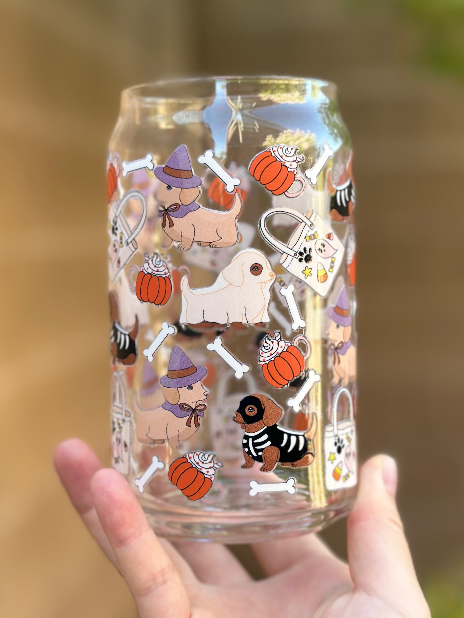 Festive glass can cup with multiple dachshunds in Halloween costumes, pumpkins, candy mugs, tote bags, and white bones on a clear background.