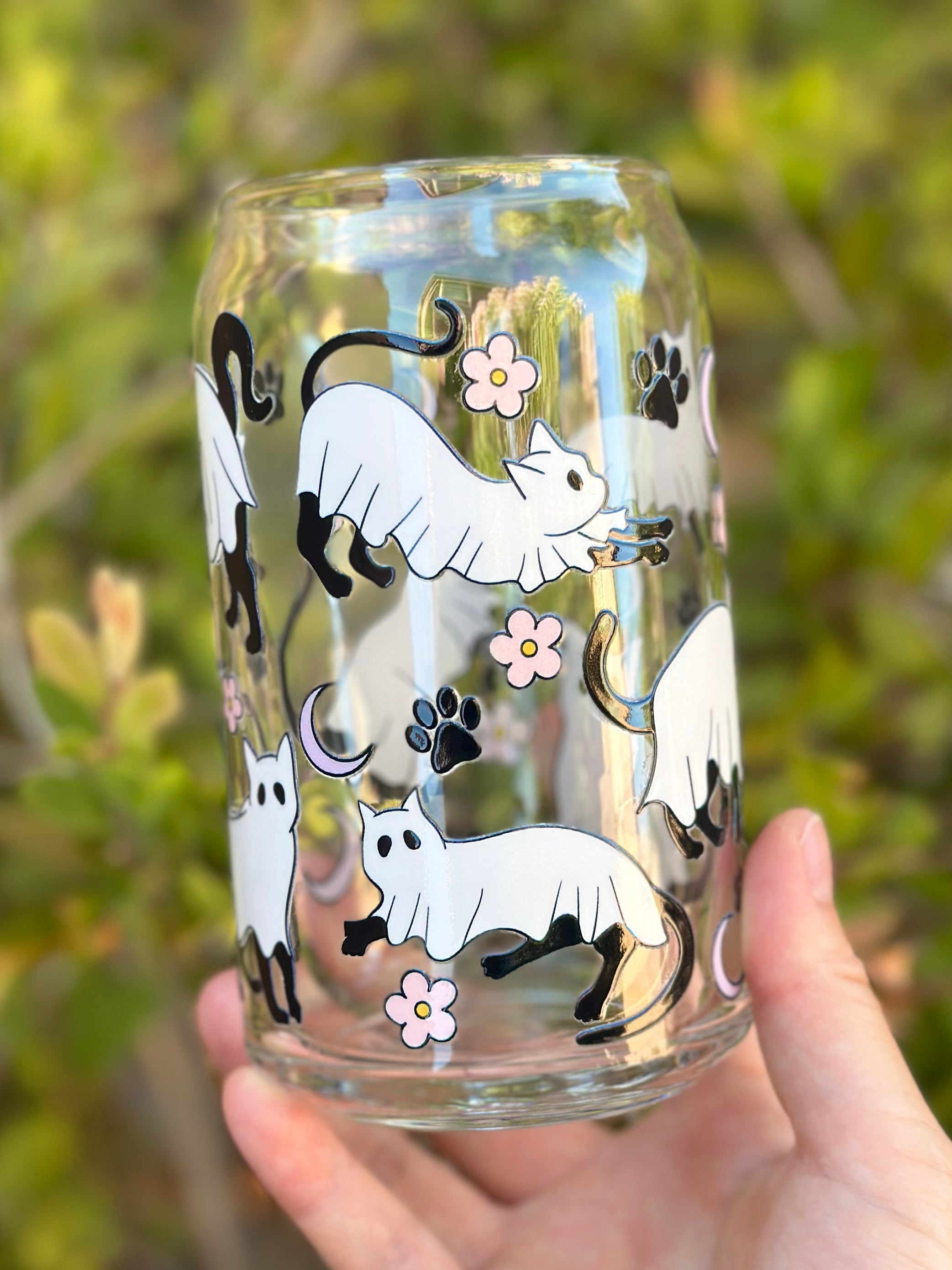 Glass can cup with black cats in ghost costumes, stretching and walking among pink flowers, paw prints, and moons.