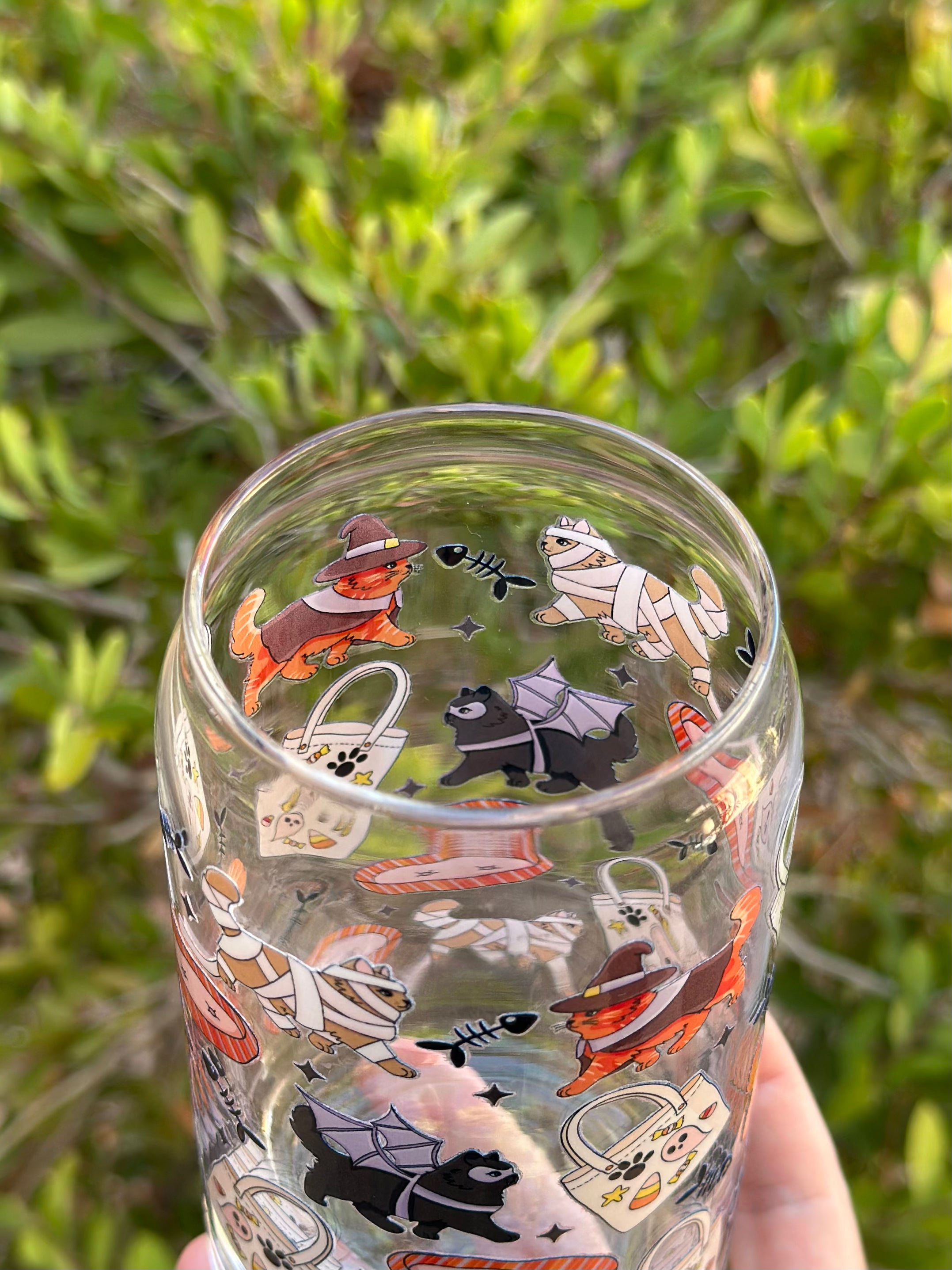 Close-up of Halloween-themed glass can cup with whimsical cats and dogs in witch, mummy, and bat costumes, with candy tote bags and fishbone designs.