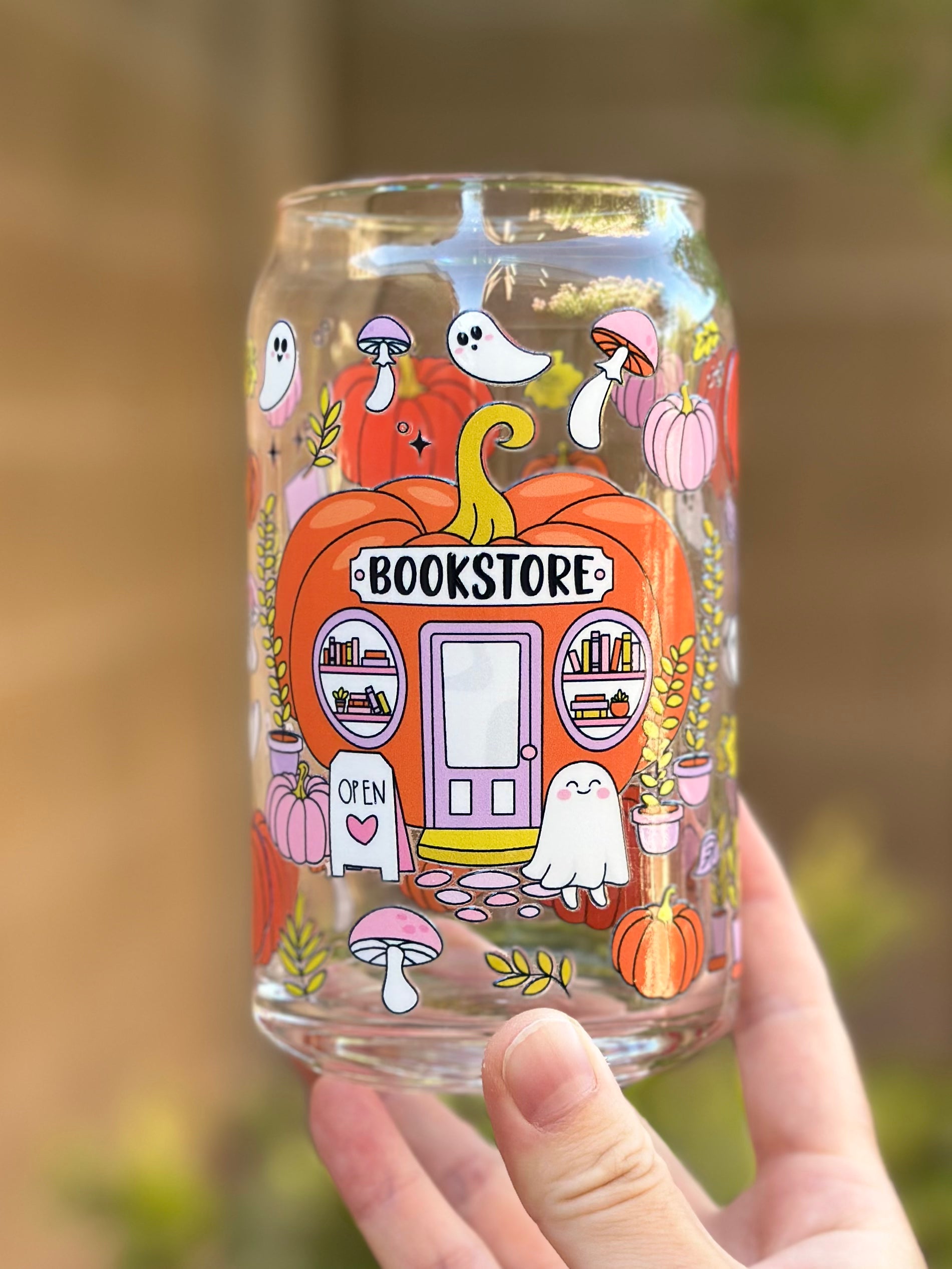 Clear glass can cup with a cute pumpkin-shaped bookstore design, surrounded by ghosts, mushrooms, pumpkins, and plants in pastel fall colors.