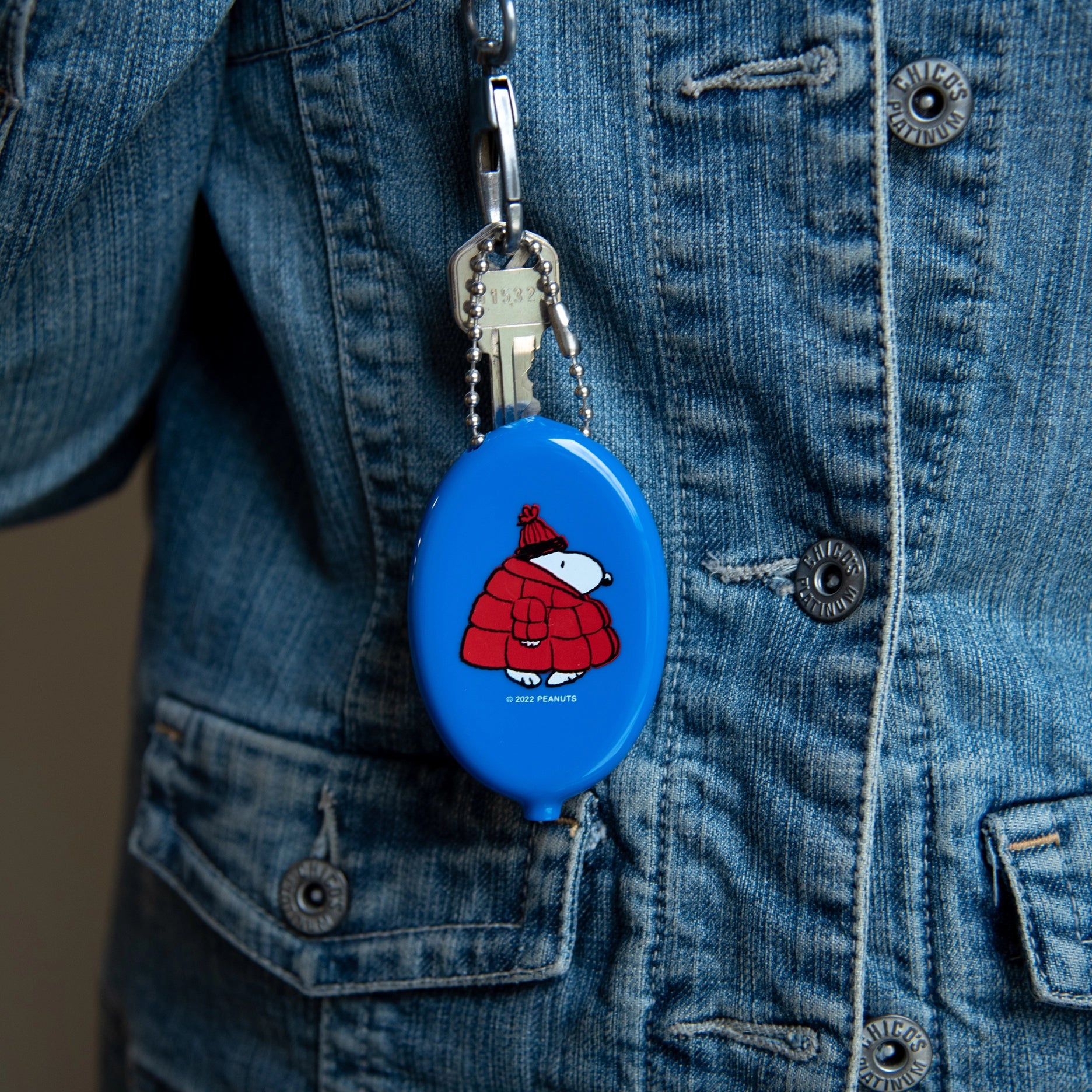 Snoopy coin pouch keychain clipped to a denim jacket. Features Snoopy in a red puffer coat and beanie, attached with a silver ball chain and key — a cozy winter-ready Peanuts accessory.