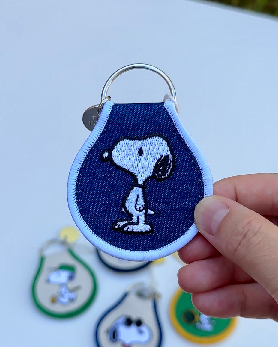Close-up of embroidered Snoopy keychain on denim blue fabric with white trim, held in hand over blurred keychains in the background.