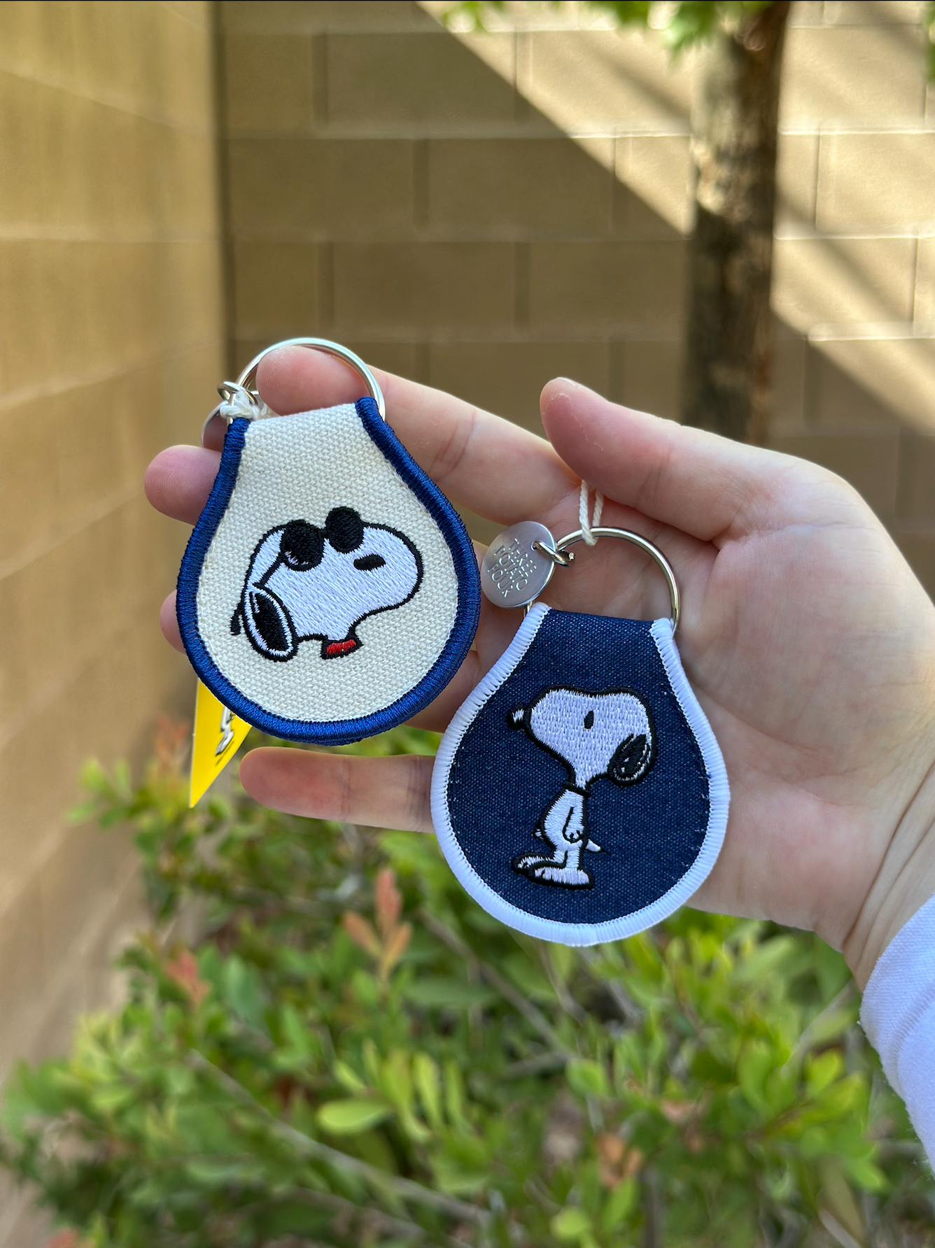 Hand holding two embroidered Snoopy keychains – one with Snoopy in sunglasses on cream fabric, and one classic standing Snoopy on denim blue with white trim.