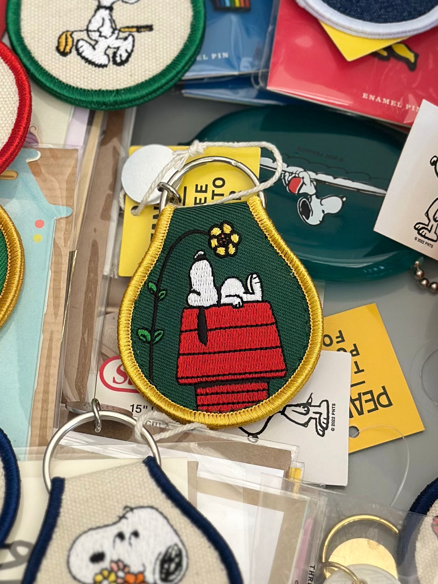 Embroidered Snoopy keychain lying on red doghouse with yellow flower, green fabric and yellow trim.