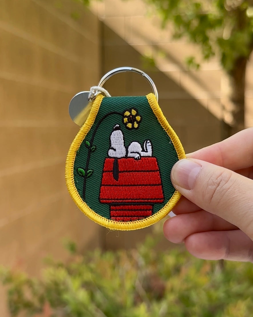 Close-up of embroidered Snoopy keychain with red doghouse, yellow flower, and green fabric background, held in hand.