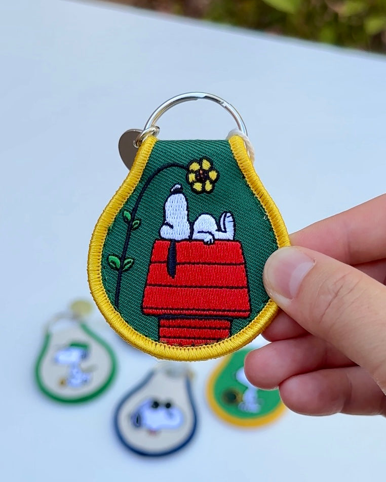 Embroidered Snoopy keychain featuring Snoopy resting on a red doghouse with a yellow flower detail, green background, and yellow trim.