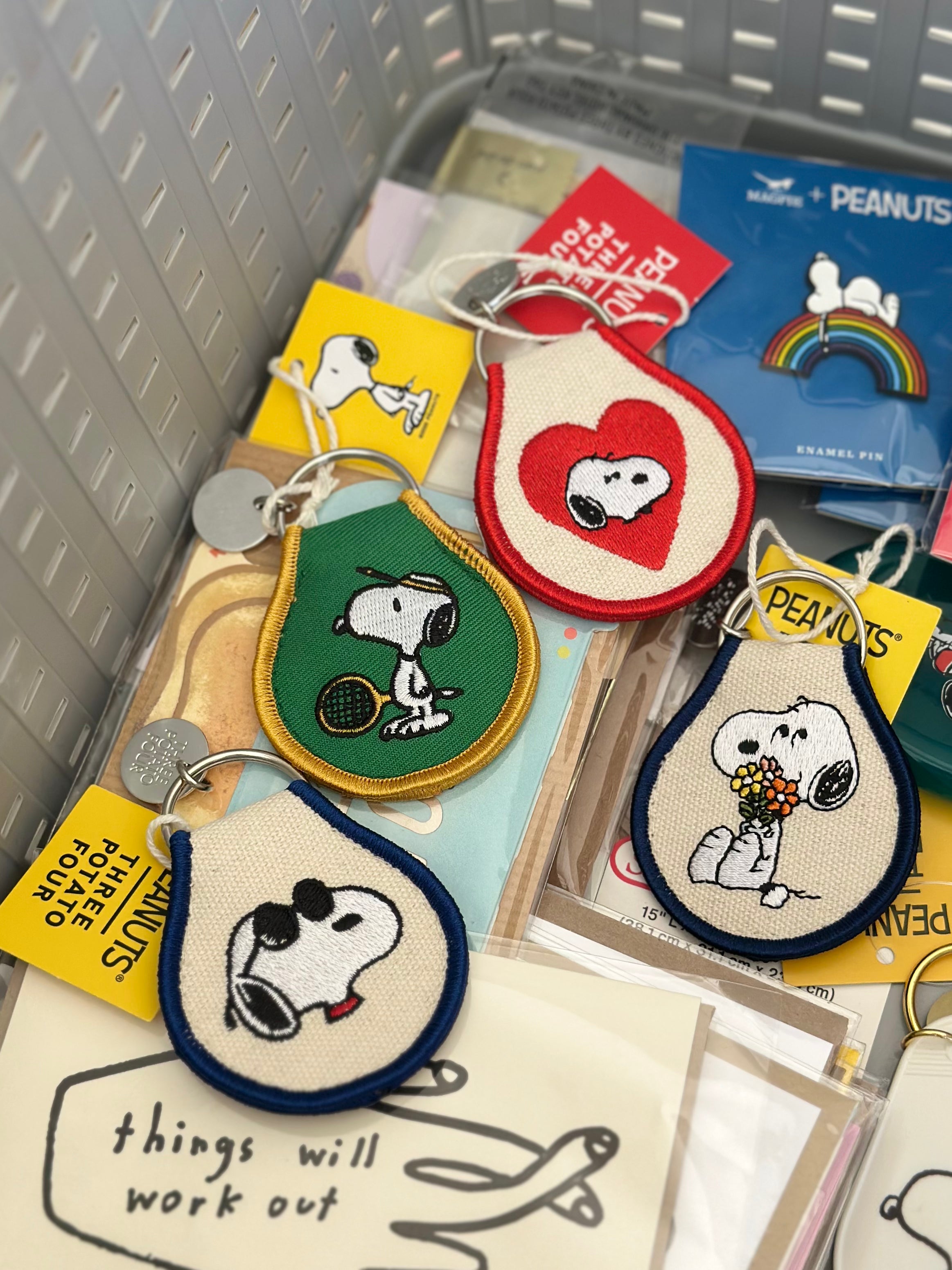 Assorted embroidered Snoopy keychains with tennis, flowers, heart, and sunglasses designs in a basket.