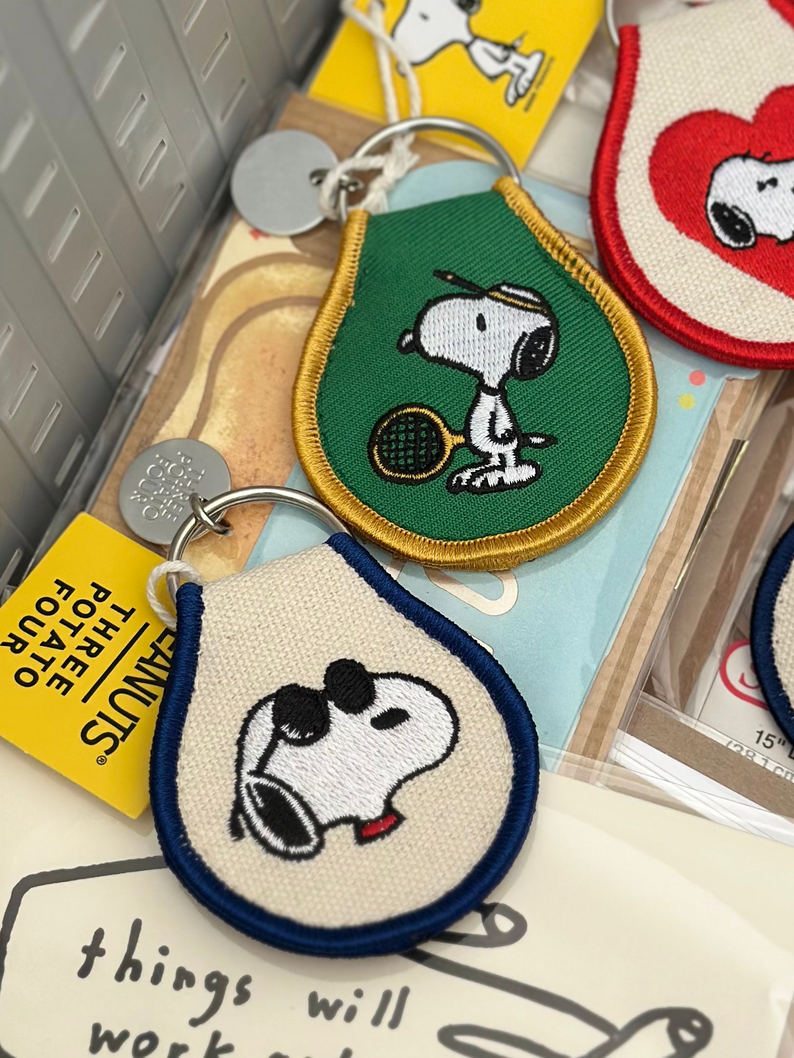 Close-up of Snoopy embroidered keychains featuring sunglasses design on cream canvas and tennis design on green canvas.