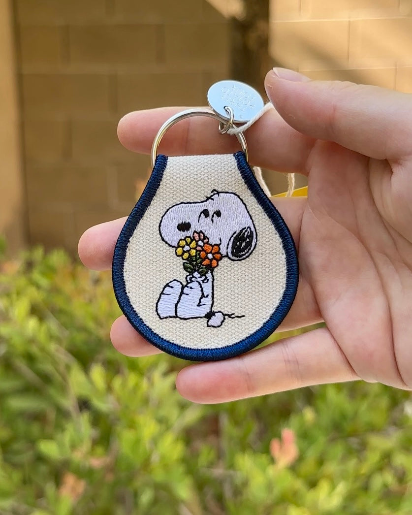 Hand holding an embroidered Snoopy keychain with Snoopy carrying a colorful flower bouquet, outlined in navy blue, official Peanuts® merch.
