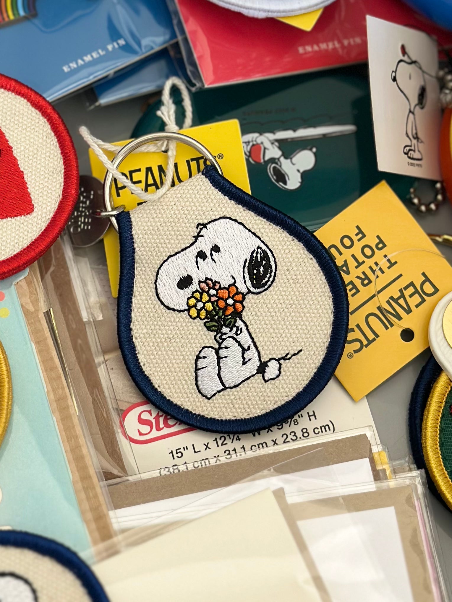 Embroidered Snoopy keychain holding bouquet of colorful flowers on cream canvas with navy trim.