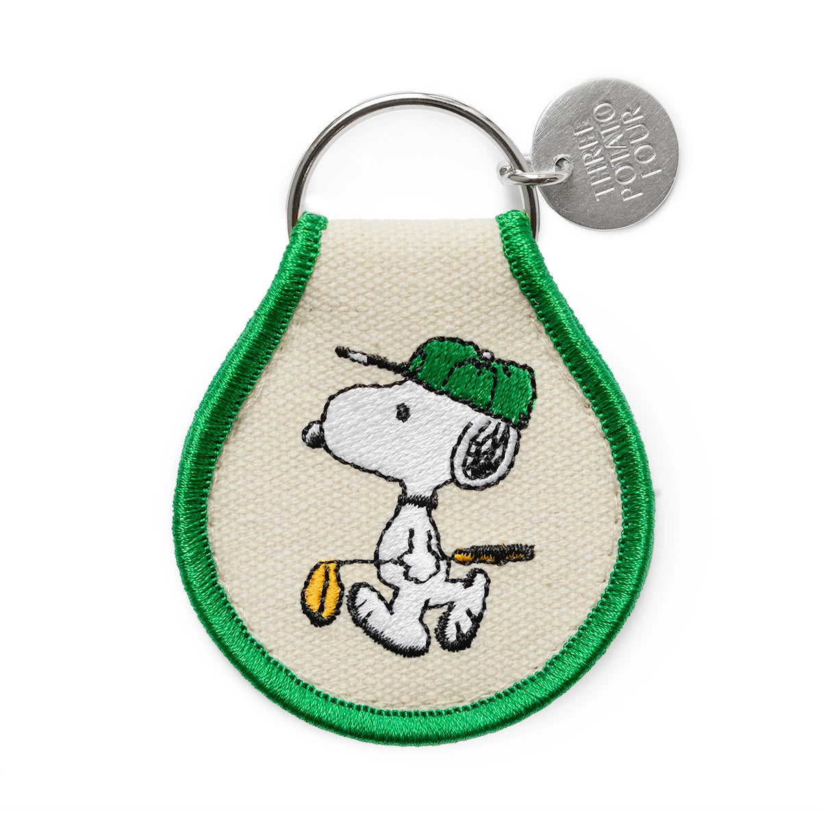 Close-up of embroidered Snoopy golf keychain with canvas texture, green border, and metal Three Potato Four charm. Features Snoopy in a green cap holding a yellow golf club — a playful and sporty Peanuts collectible.