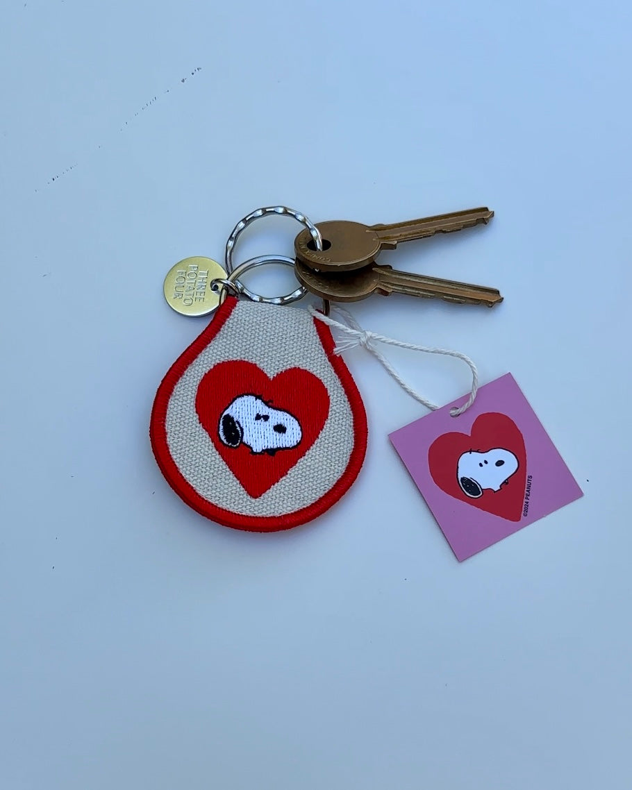 Close-up of Snoopy Heart embroidered keychain with red heart design, attached to gold keys and pink Peanuts tag.
