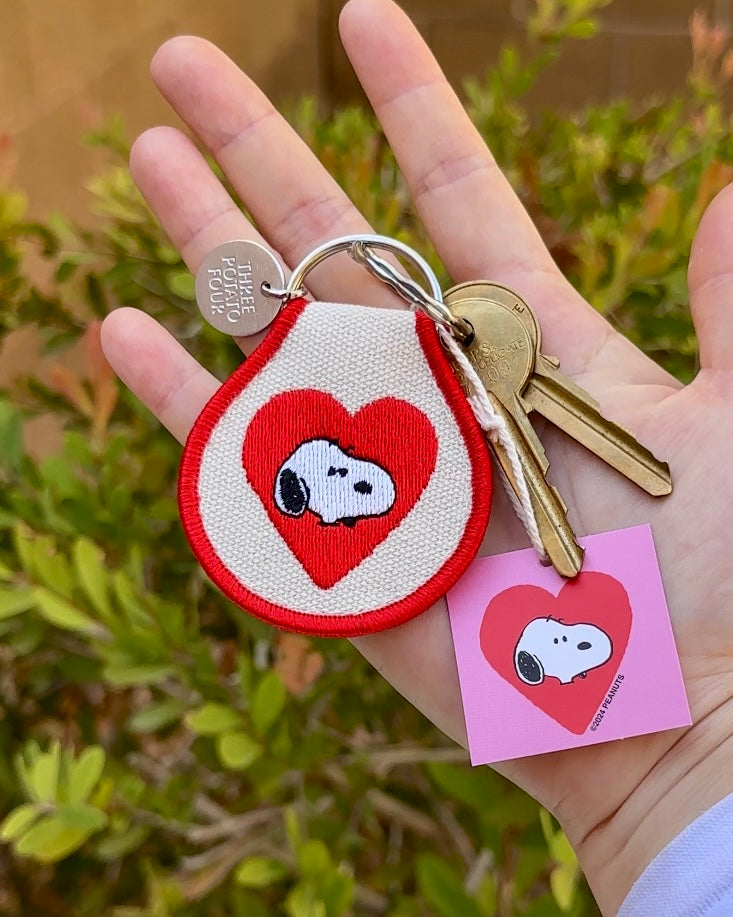 Snoopy Heart embroidered patch keychain with gold keys, photographed outdoors with greenery background.