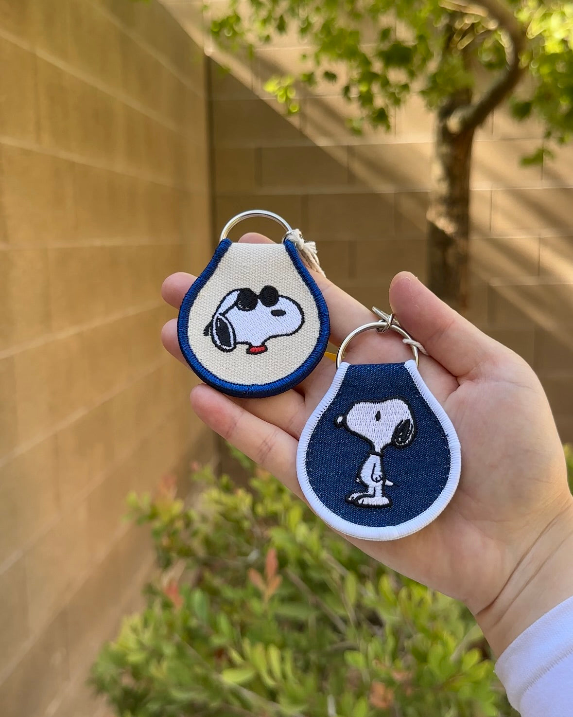 Two Snoopy Peanuts embroidered keychains in blue and white trim, featuring sunglasses Snoopy and standing Snoopy, held outdoors.