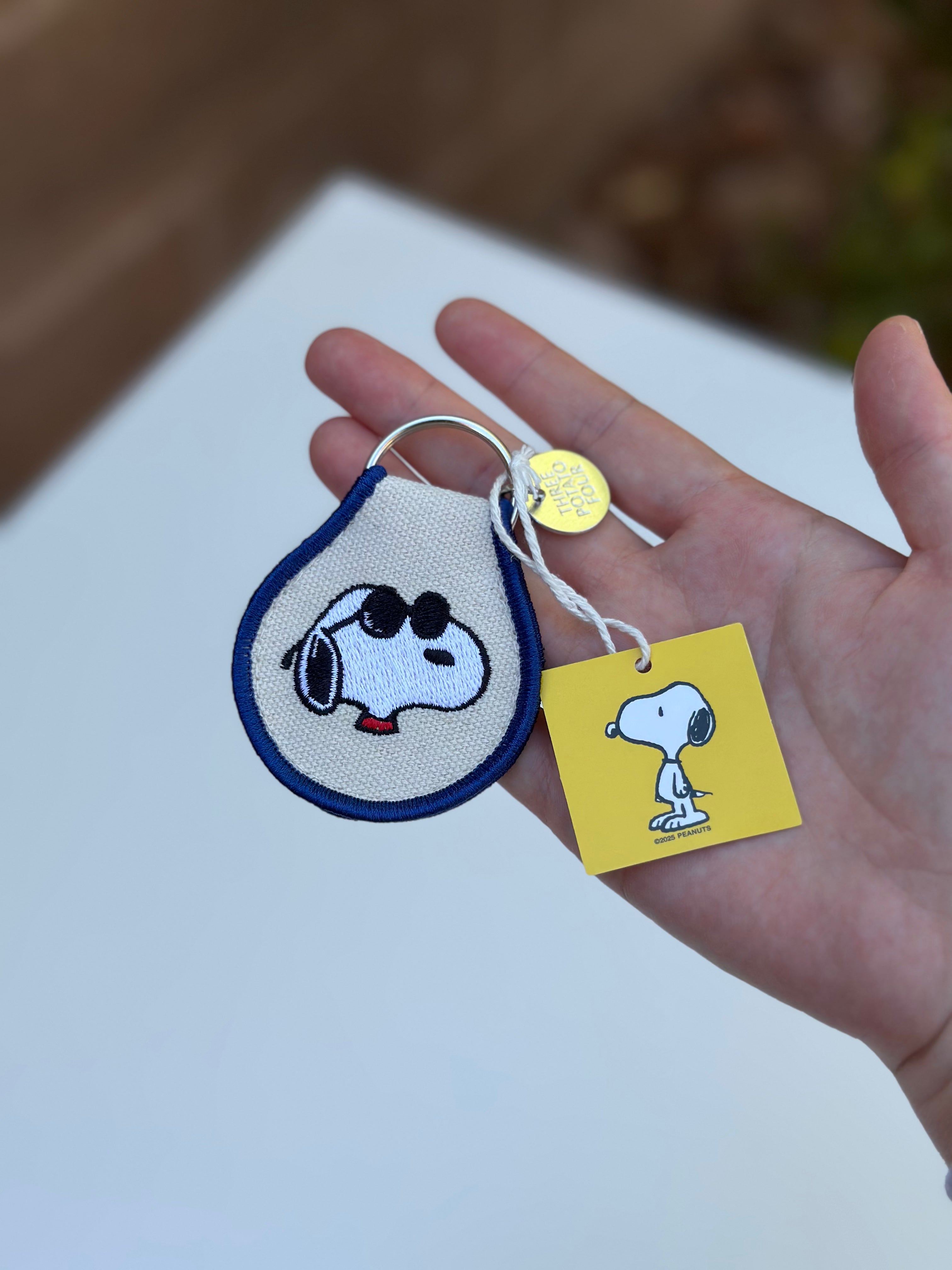 Snoopy Peanuts embroidered keychain with sunglasses design and blue trim, held in hand with yellow Peanuts tag visible.