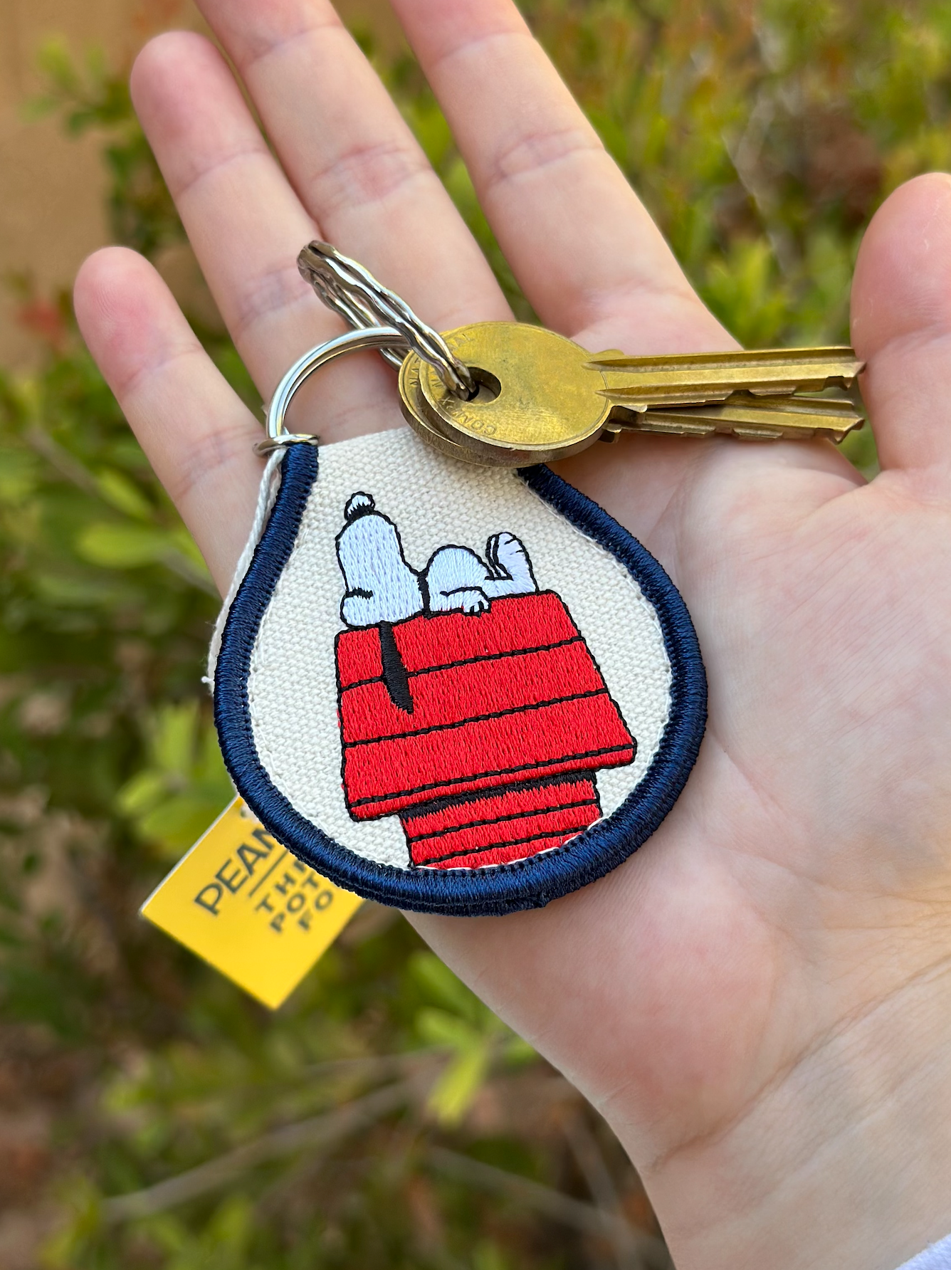 Hand holding Snoopy Peanuts embroidered keychain with Snoopy sleeping on his red doghouse, navy trim, and silver split ring.
