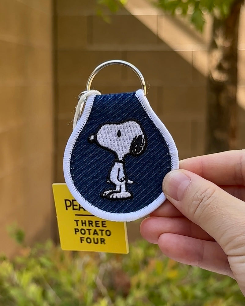 Embroidered Snoopy keychain featuring standing Snoopy on denim blue background with white trim, held in hand outdoors.
