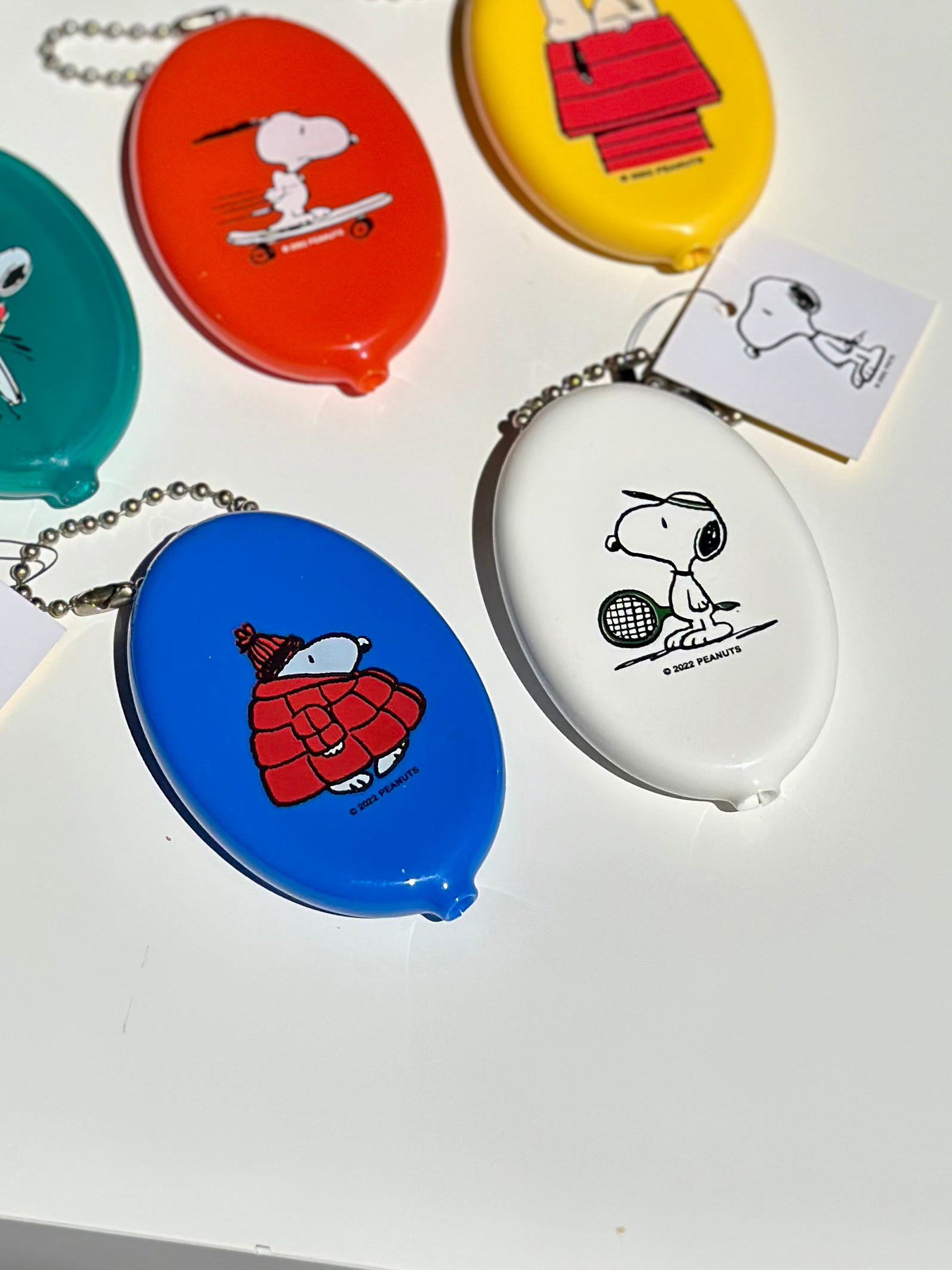 White Snoopy tennis coin pouch with ball chain keyring, shown with other colorful Peanuts® pouches.