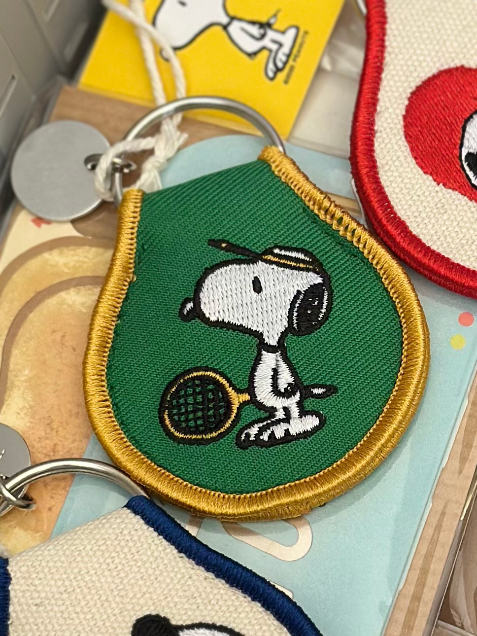 Embroidered Snoopy tennis keychain on green canvas with gold trim and metal keyring.