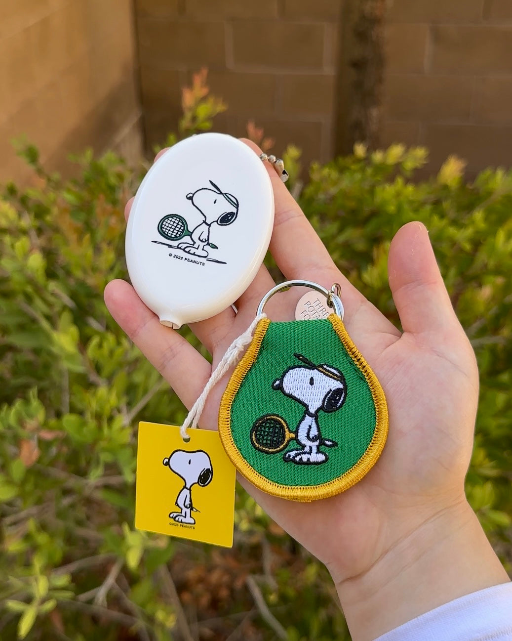 Snoopy Peanuts embroidered tennis keychain with yellow trim, shown with matching white Snoopy tennis coin pouch, held outdoors.