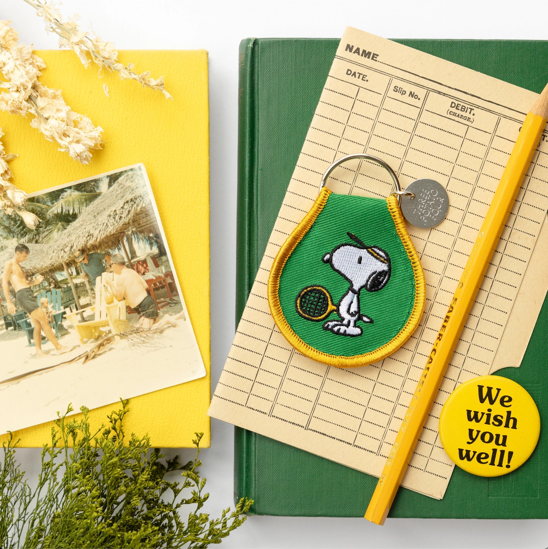 Flat lay featuring a Snoopy tennis keychain on top of retro library cards, books, and a yellow pencil. Embroidered green and yellow keychain with vintage charm — a nostalgic Peanuts-inspired gift or accessory.