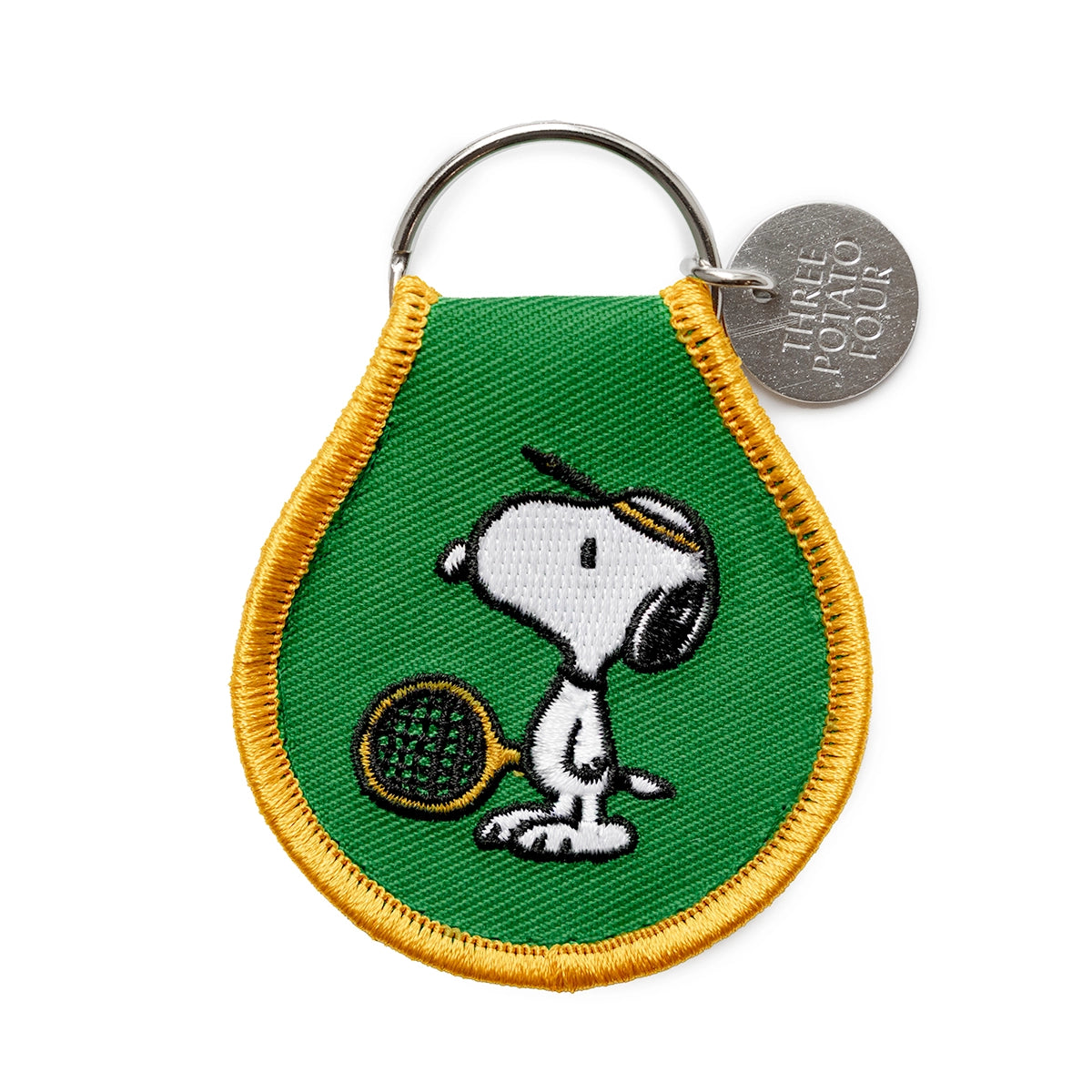 Close-up of embroidered Snoopy keychain with green background and yellow trim. Features Snoopy in a tennis visor holding a racket, attached to a silver key ring with Three Potato Four branding tag.