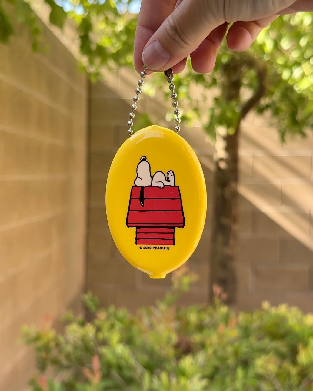 Close-up of yellow Snoopy coin pouch featuring Peanuts® Snoopy napping on red doghouse, retro squeeze wallet style with ball chain keyring.