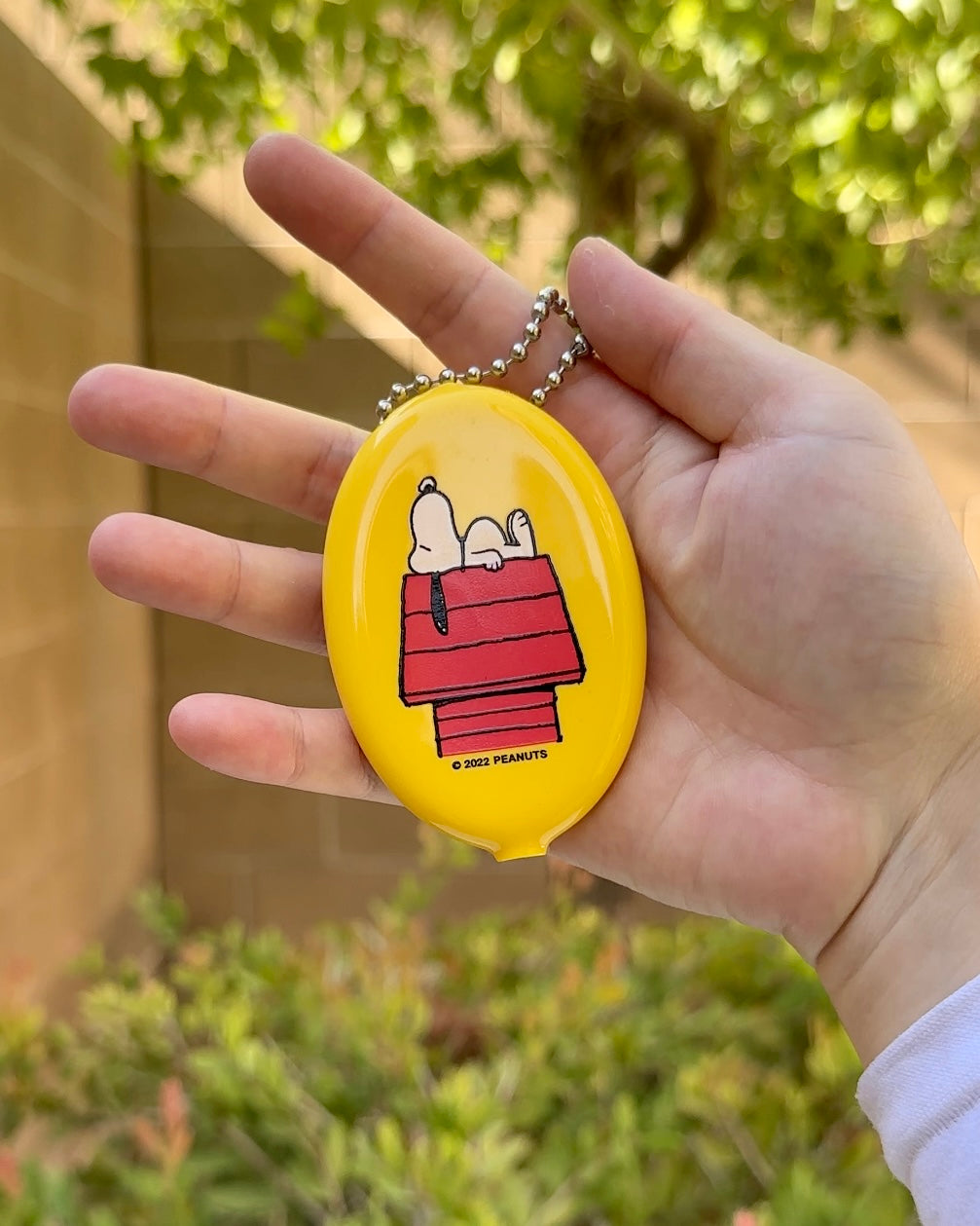 Yellow Peanuts® Snoopy vinyl coin pouch with squeeze-to-open design, showing Snoopy sleeping on red doghouse, ball chain keyring for bags or keys.