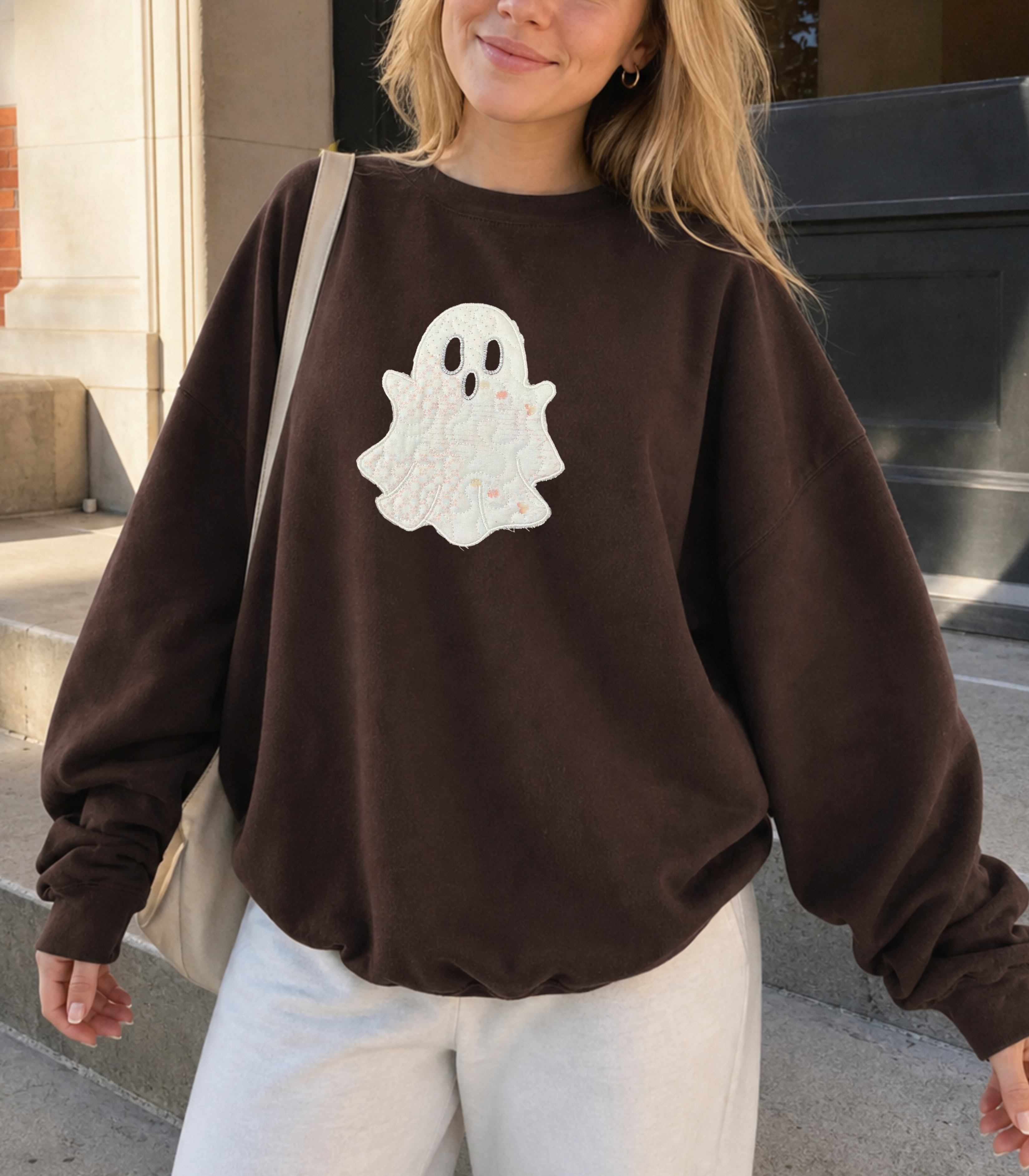 A woman wearing a dark brown crewneck sweatshirt with a white ghost applique on the front, paired with off-white pants and carrying a beige shoulder bag, standing outdoors on concrete steps.