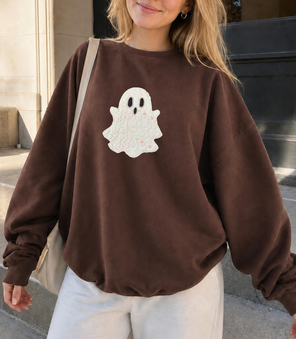 A woman wearing a brown crewneck sweatshirt with a white ghost applique on the front, paired with white sweatpants and a beige shoulder bag, standing outdoors on concrete steps.