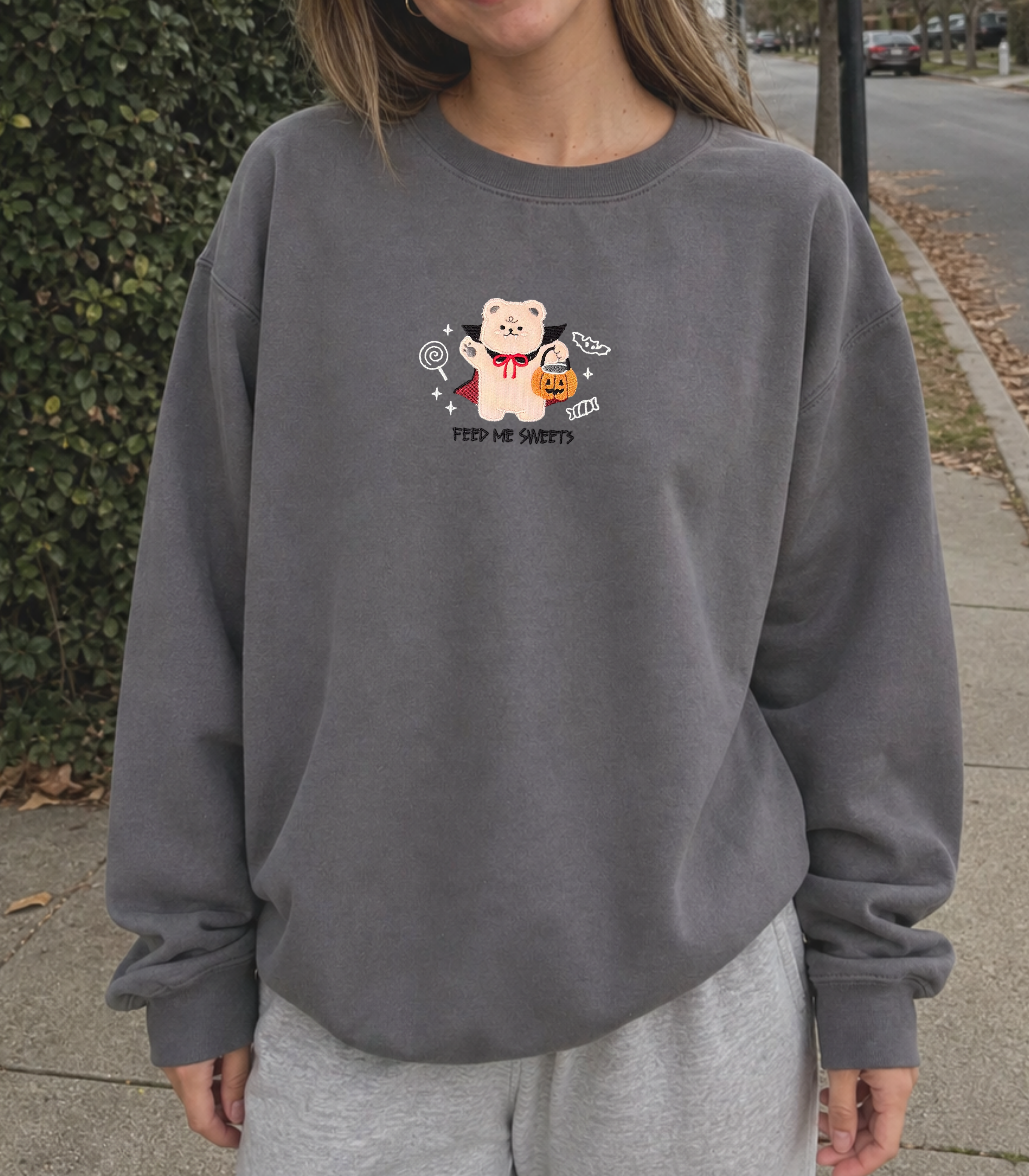 A person wearing a dark gray crewneck sweatshirt with a cute cartoon bear graphic holding a pumpkin and tennis rackets, standing outdoors on a sidewalk.