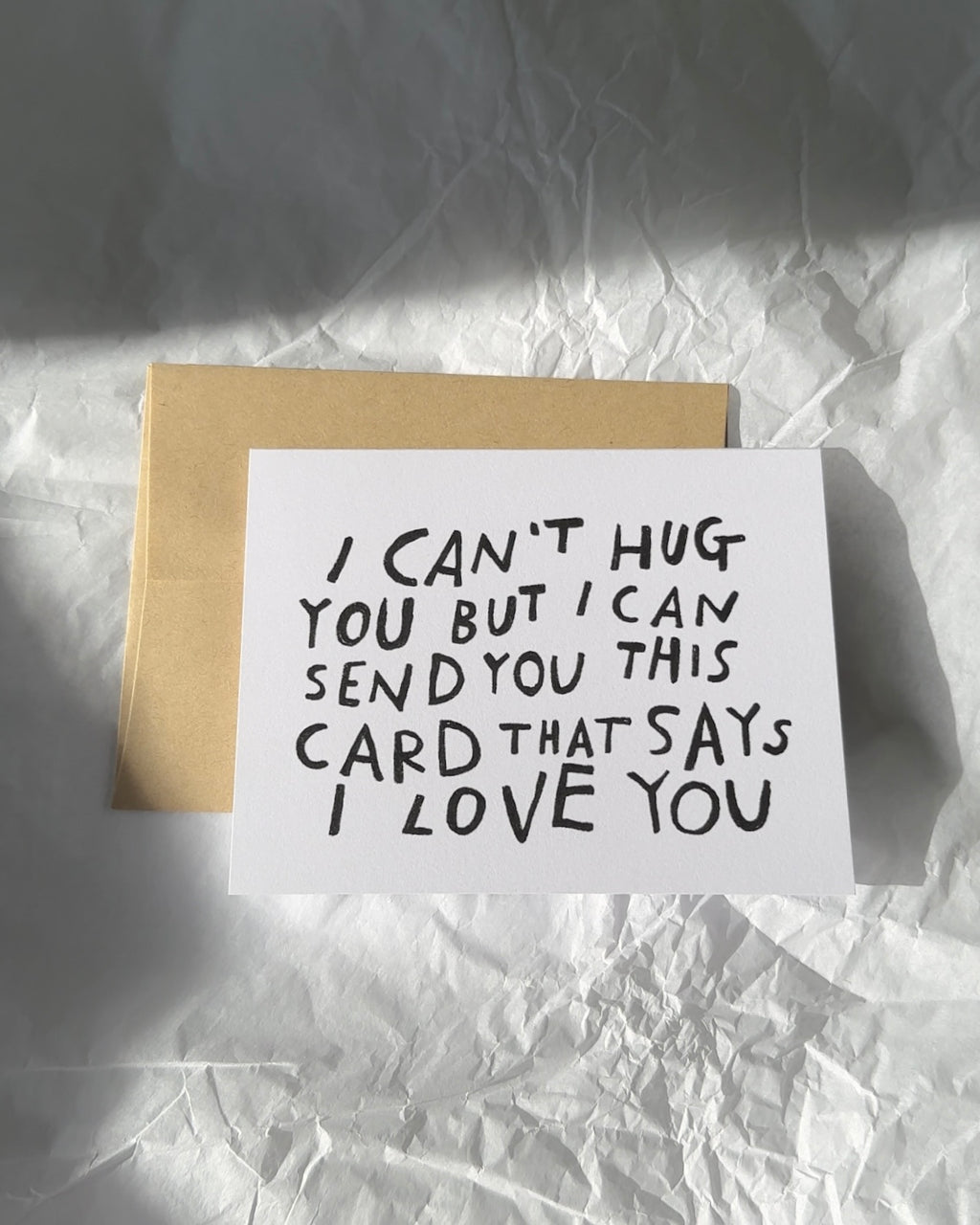 Minimalist white greeting card with black hand-lettered text that reads ‘I can’t hug you but I can send you this card that says I love you,’ paired with brown kraft envelope.