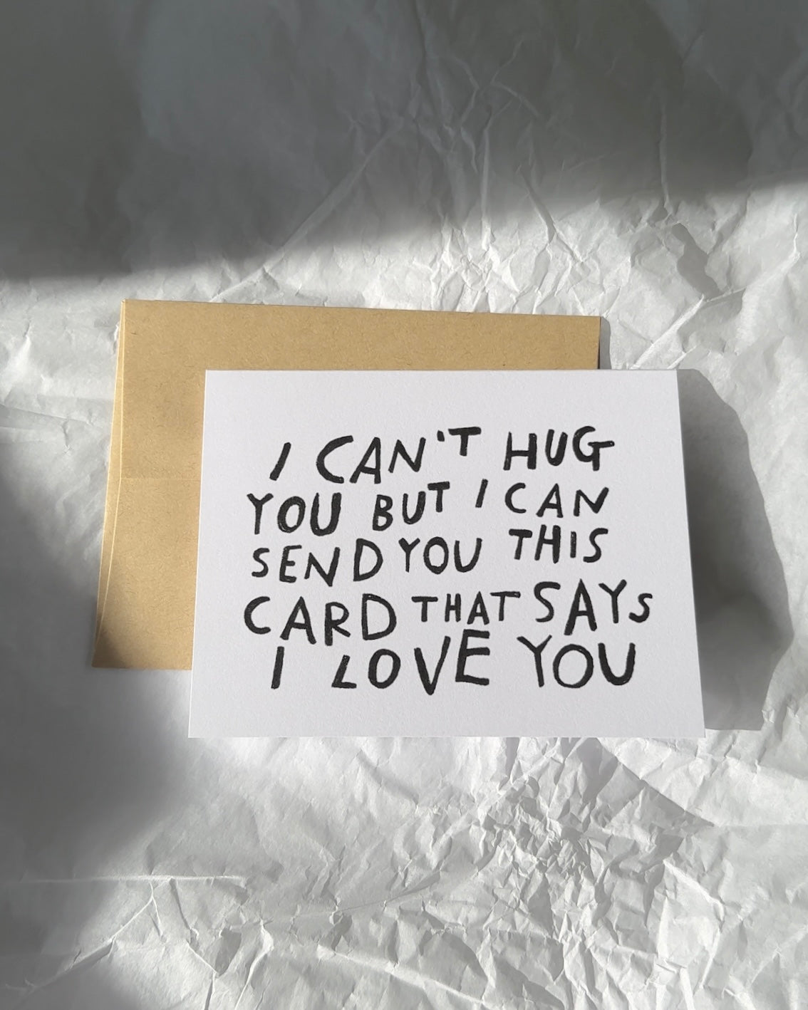 Minimalist white greeting card with black hand-lettered text that reads ‘I can’t hug you but I can send you this card that says I love you,’ paired with brown kraft envelope.