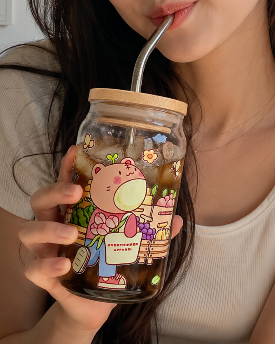 A close-up of a person holding a clear glass jar tumbler with a bamboo lid and metal straw, decorated with a cute cartoon bear illustration. The person is wearing a beige ribbed tank top and holding the glass with one hand while drinking from it with the other hand off-frame.