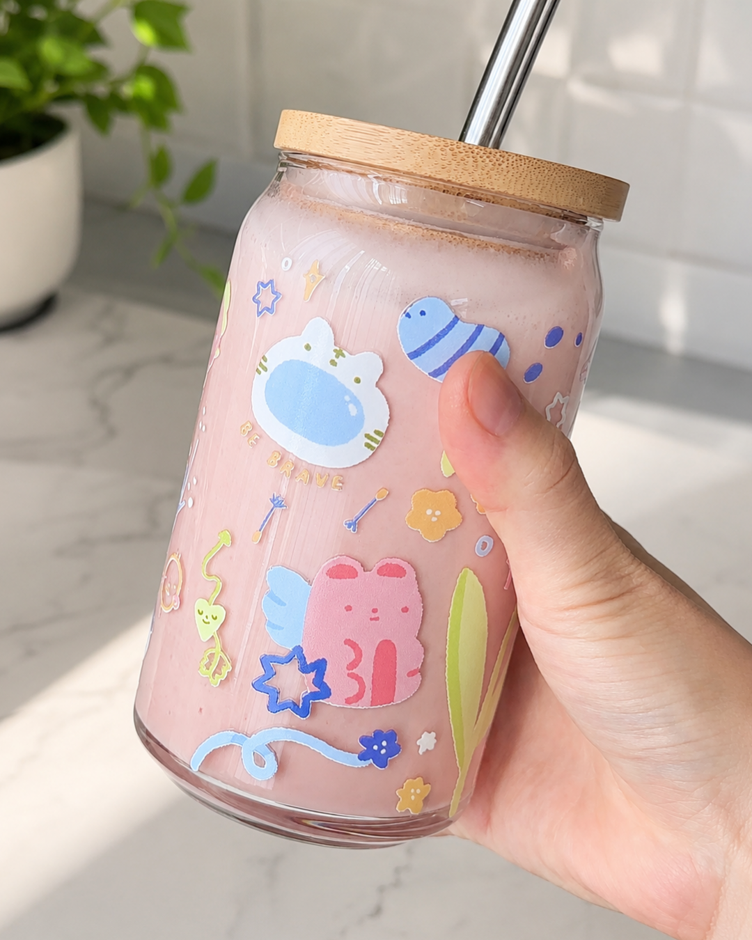 A hand holding a clear glass tumbler with a bamboo lid and metal straw, filled with a pink drink. The tumbler is decorated with cute cartoon stickers including a pink bear, a blue fish, and a white cat face.
