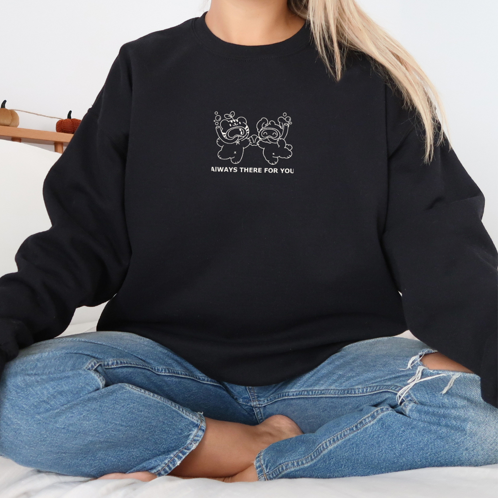 Cozy black sweatshirt with “Always There For You” bear graphic, modeled seated cross-legged in a casual lifestyle setting.