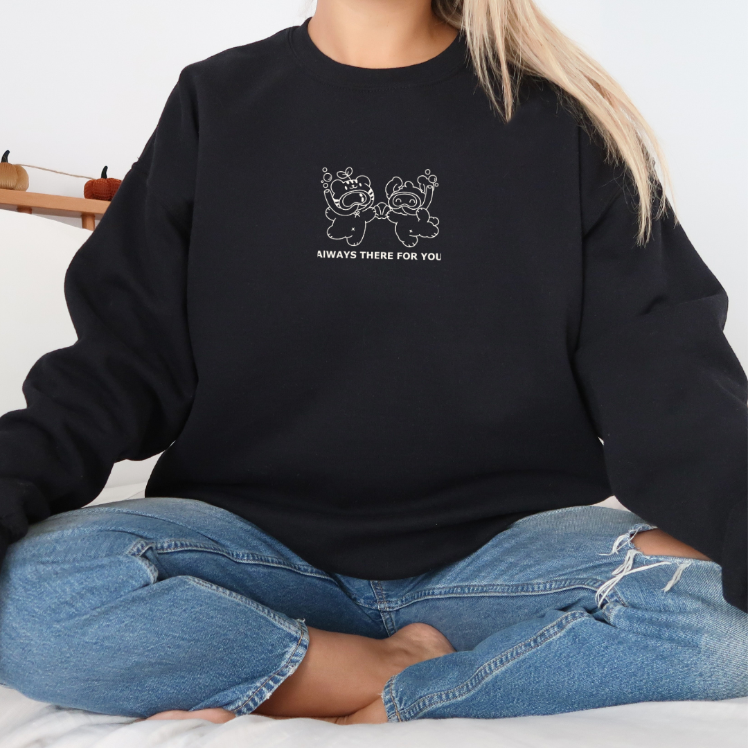 Cozy black sweatshirt with “Always There For You” bear graphic, modeled seated cross-legged in a casual lifestyle setting.