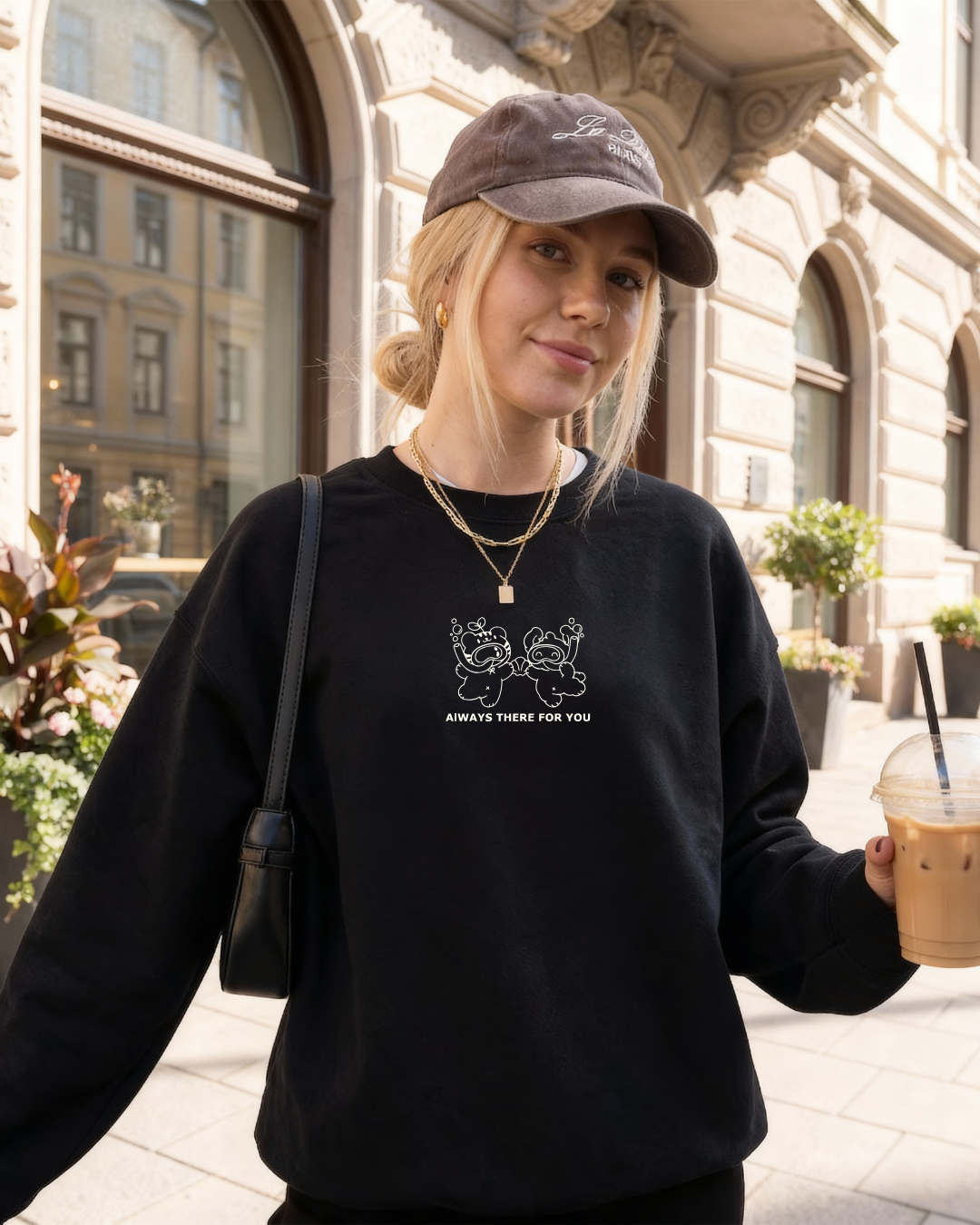 Woman wearing a black oversized embroidered crewneck sweatshirt with “always there for you” text while holding an iced coffee outdoors