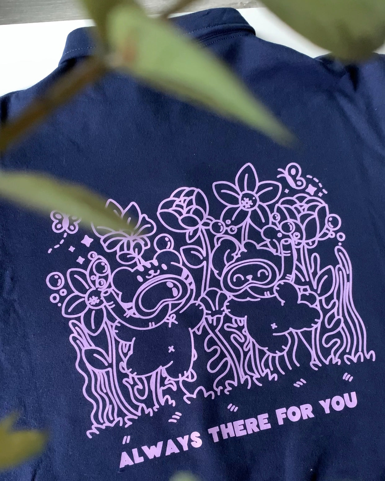 Close-up of the back illustration on a navy quarter-zip fleece pullover, showing two characters surrounded by florals with the supportive phrase “Always there for you.”