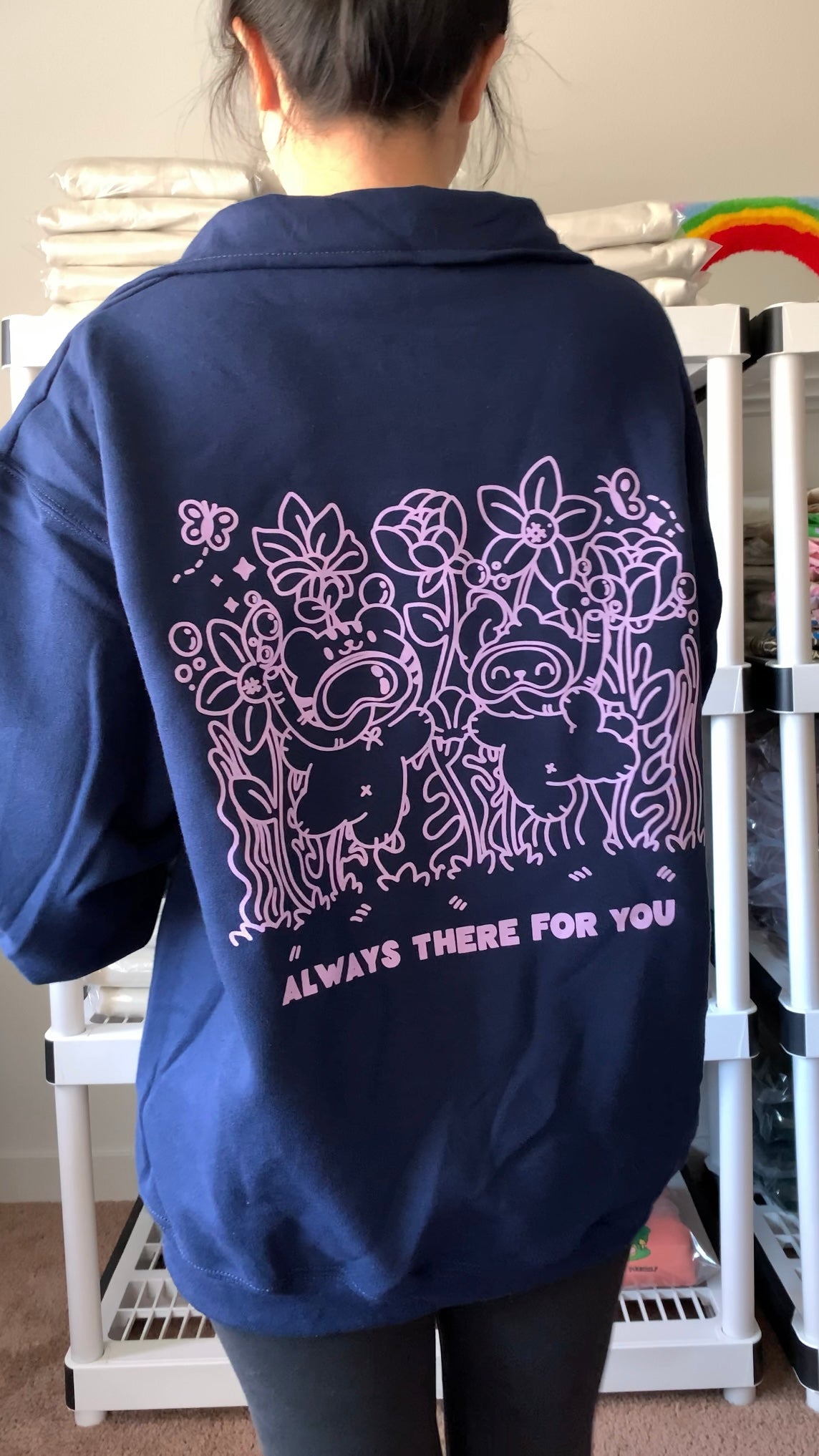 Model wearing a navy quarter-zip fleece pullover shown from the back, featuring a soft illustrated floral scene with two companions and the phrase “Always there for you,” designed to feel calm and supportive.