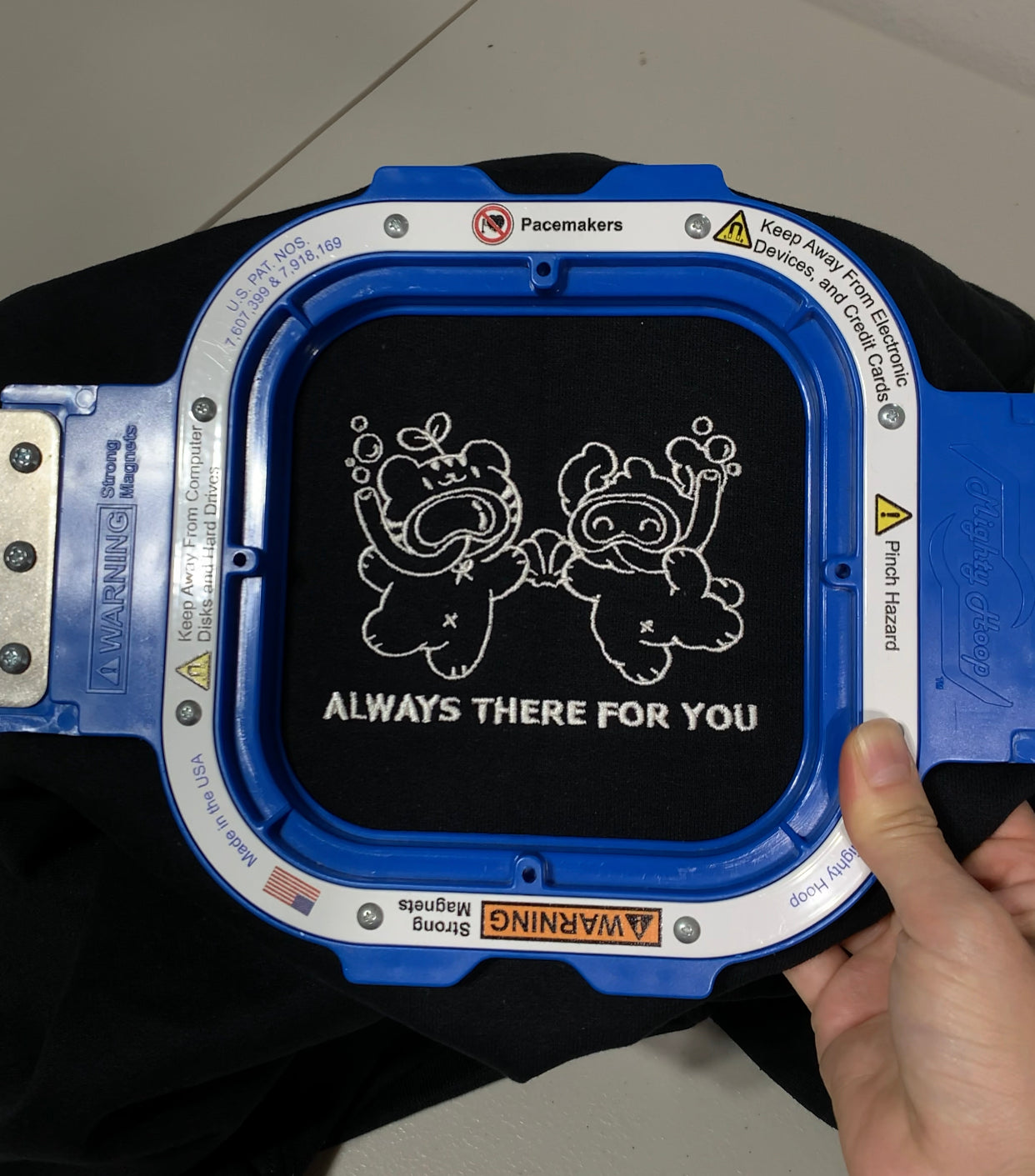Close-up of the “Always There For You” bear graphic being embroidered on a black sweatshirt using a blue embroidery hoop.