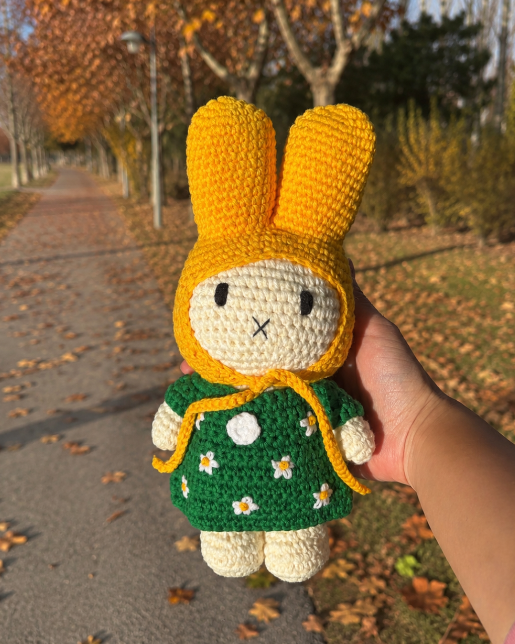 A handmade crocheted bunny doll wearing a yellow bonnet and green dress with white daisy flowers, held by a hand against an outdoor autumn park background.