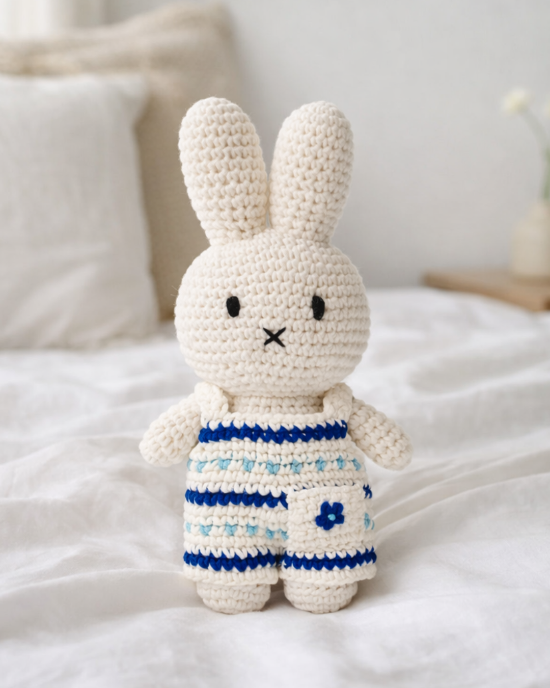 Cute amigurumi crochet Miffy plush toy in blue jumpsuit sitting on bed, handmade stuffed animal for nursery decor or baby gift idea