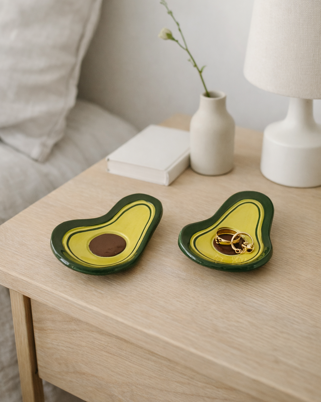 Handmade ceramic avocado ring dish on bedside table holding gold jewelry, cute fruit shaped trinket dish for nightstand decor and jewelry organization